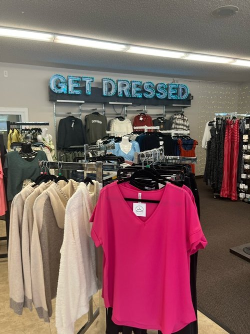 Retail clothing store display with a pink T-shirt on a hanger in the foreground, beige and gray sweaters on another rack, and a neon sign reading 'GET DRESSED' on the wall behind.