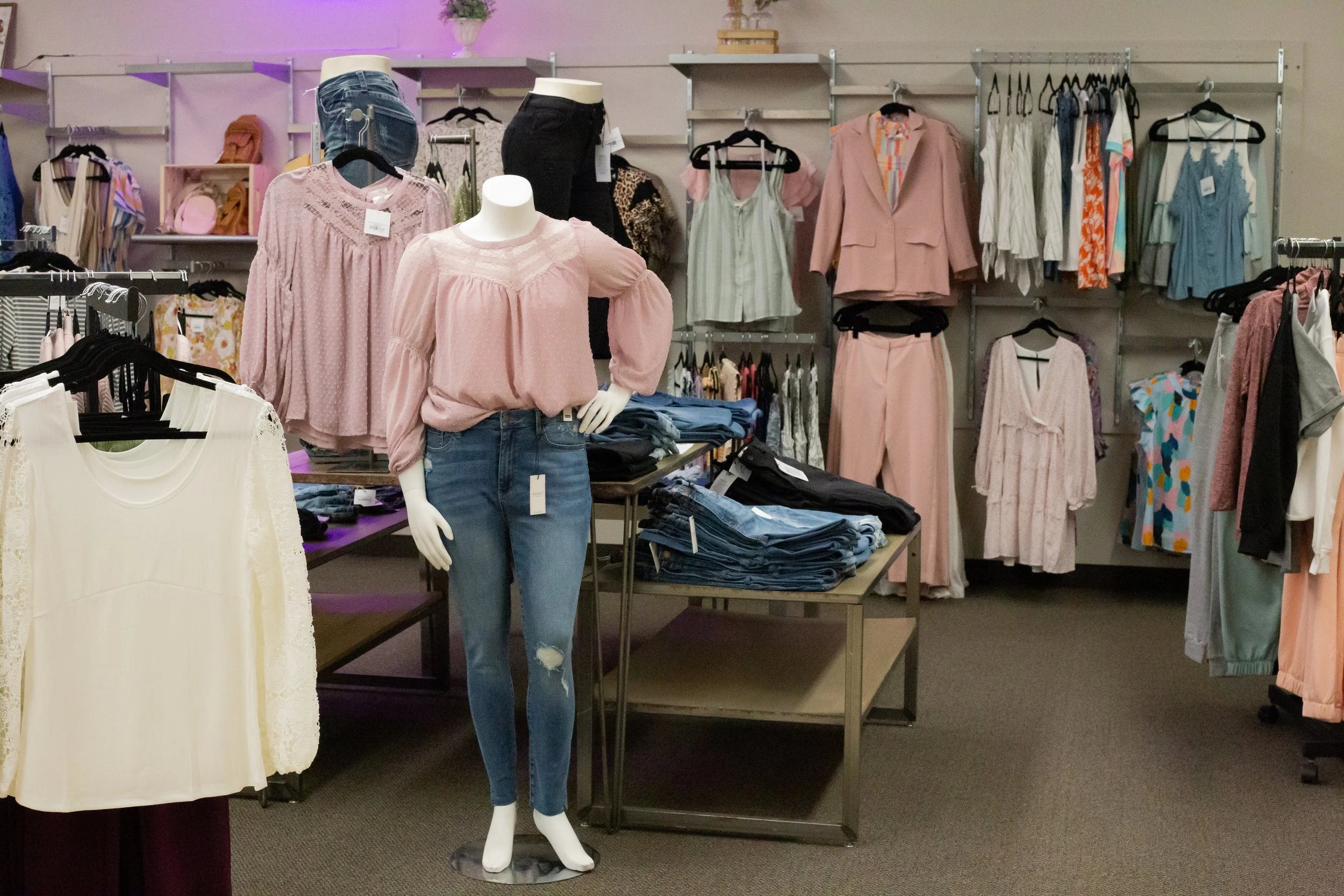 Clothing display in a retail store including a mannequin wearing a pink top and ripped jeans, and racks of various women's apparel in pastel and neutral colors.