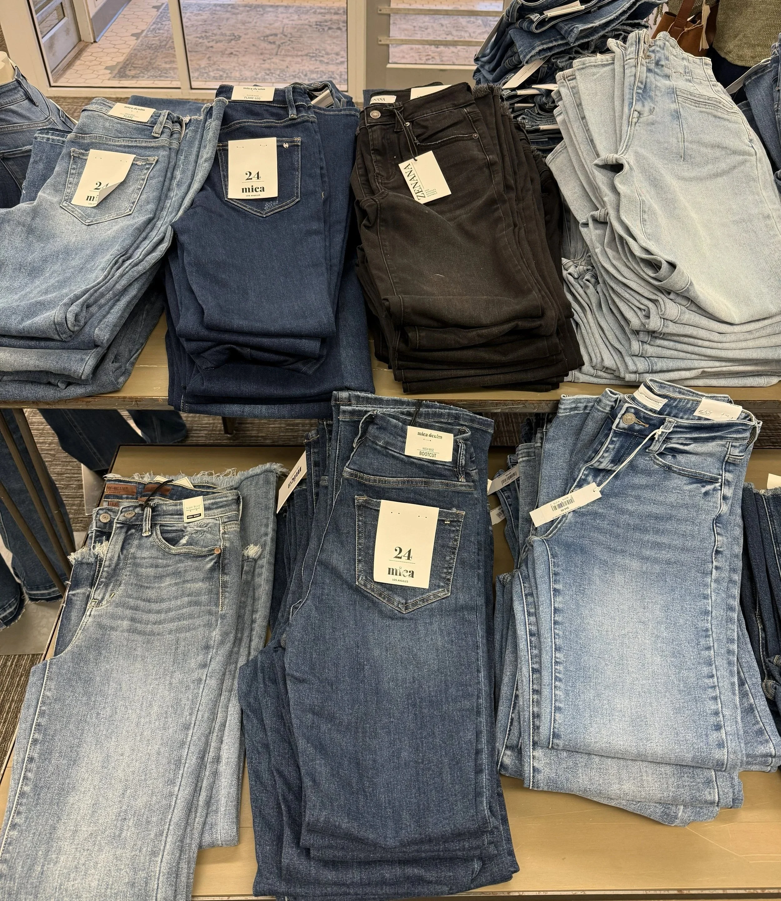 Stacked jeans in various styles and colors, including light, dark, black, and gray, displayed on tables in a retail store.