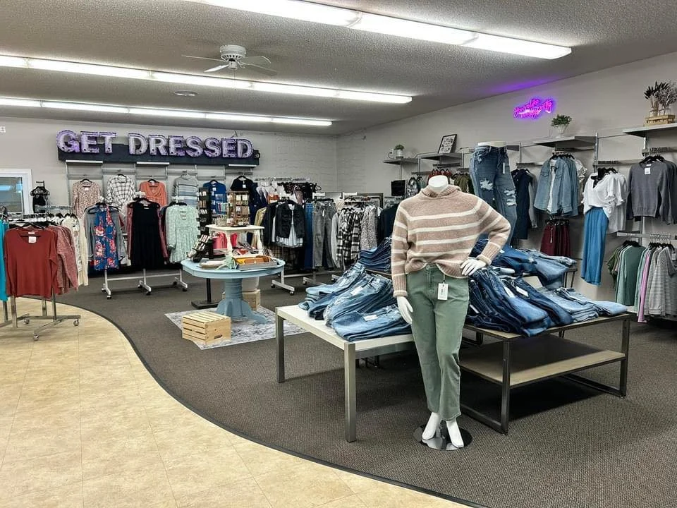 Clothing store interior with a mannequin wearing a beige striped sweater and green pants, racks of clothes including jeans, shirts, and jackets, and a neon sign that reads 'GET DRESSED'