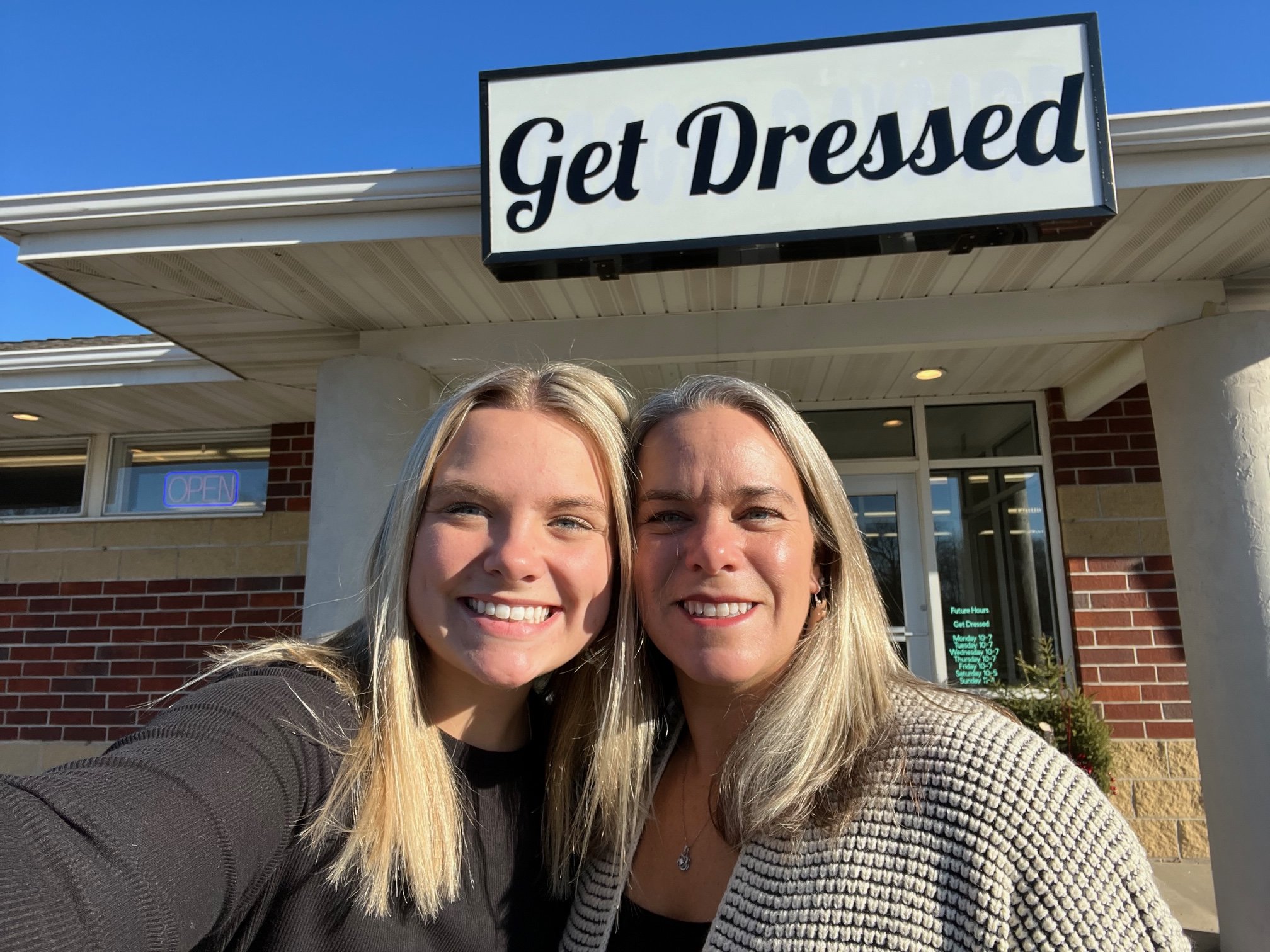Two women taking a selfie outside a store named 'Get Dressed' under a sign, during daytime with clear blue skies.