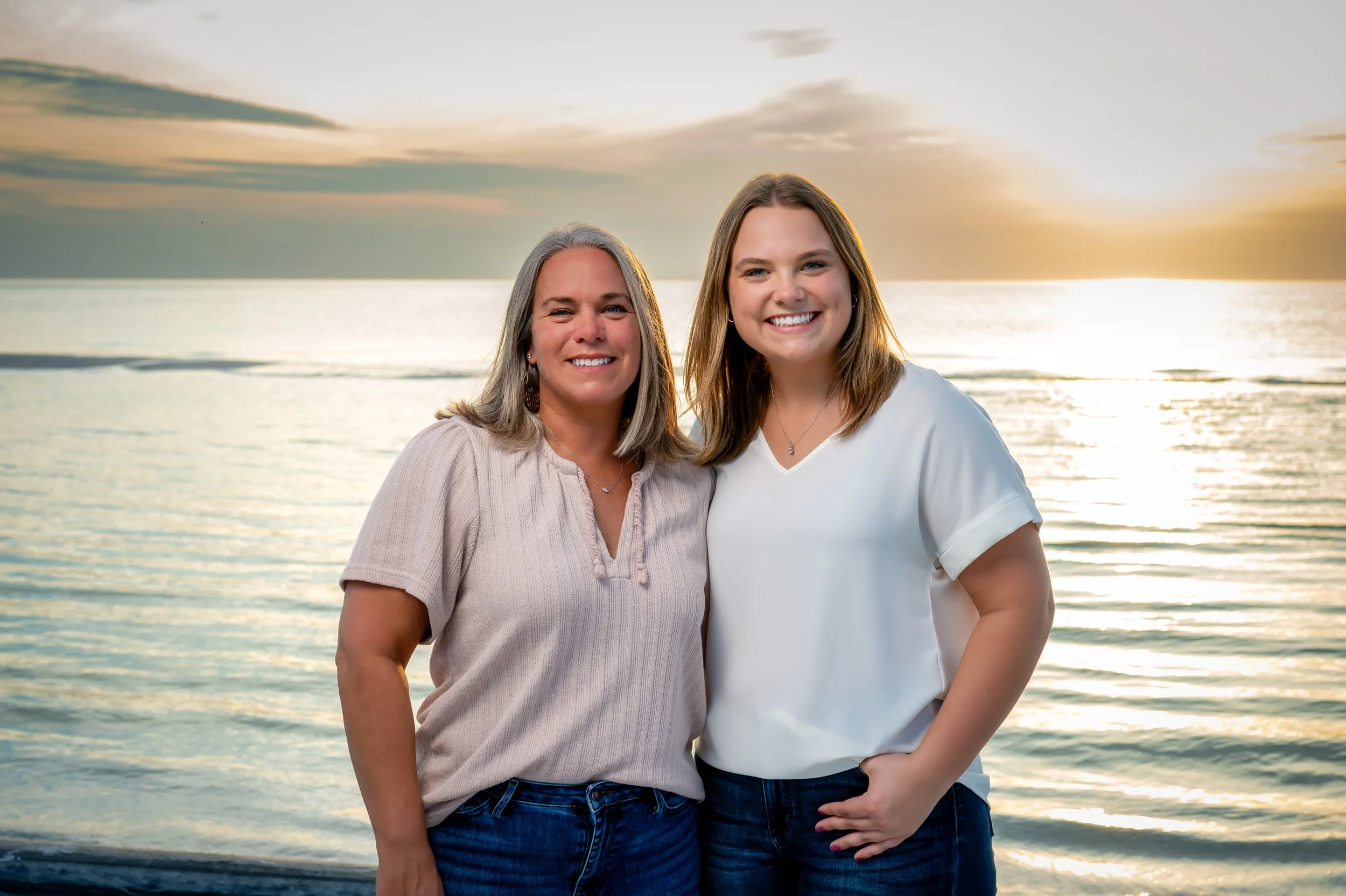 Two smiling women standing together on a beach at sunset, with calm water and cloudy sky in the background.