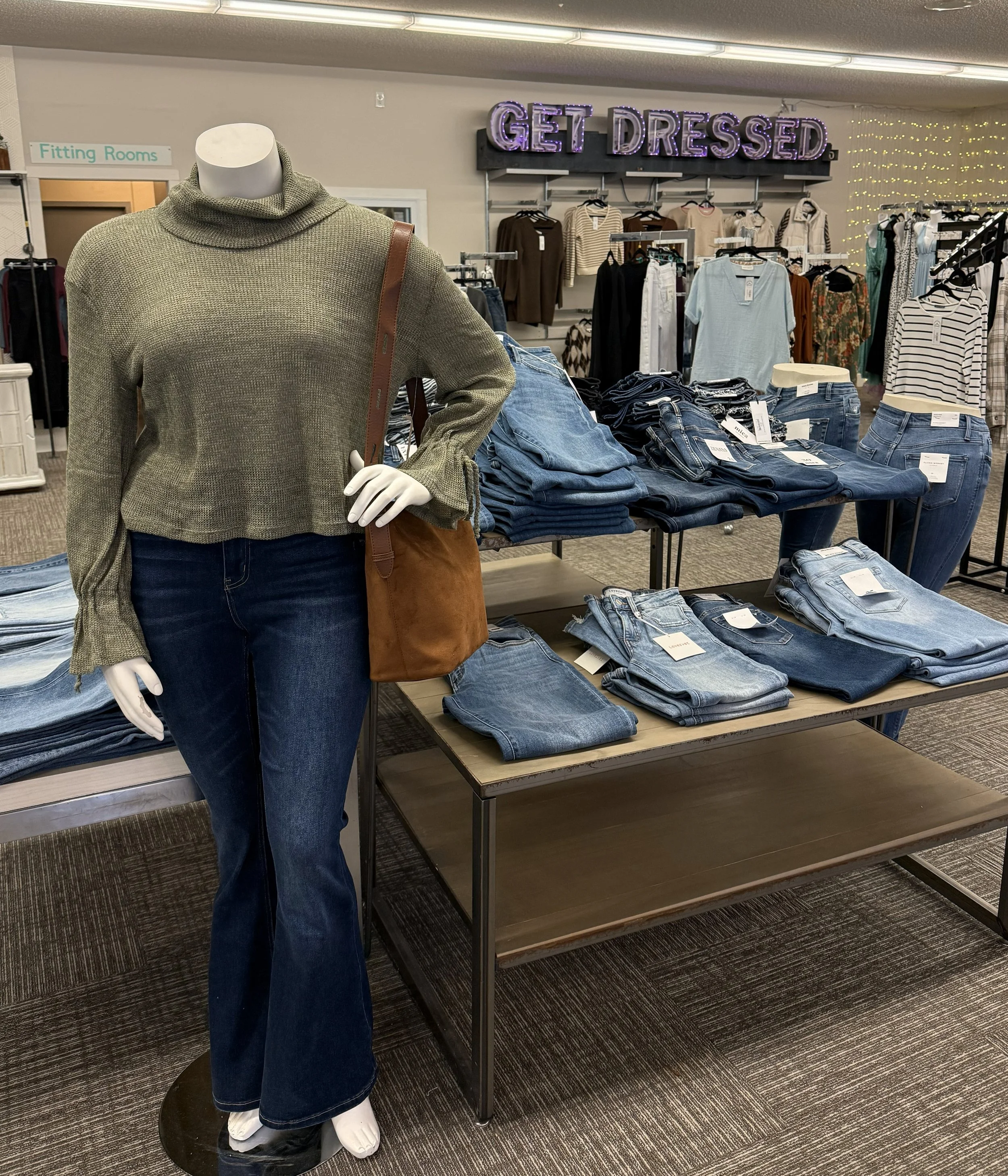 A retail store display with a mannequin wearing a green sweater, dark blue jeans, and a brown shoulder bag. Behind the mannequin, there are tables with folded jeans and a sign that says 'GET DRESSED.' In the background, clothing racks, a fitting room sign, and various garments are visible.