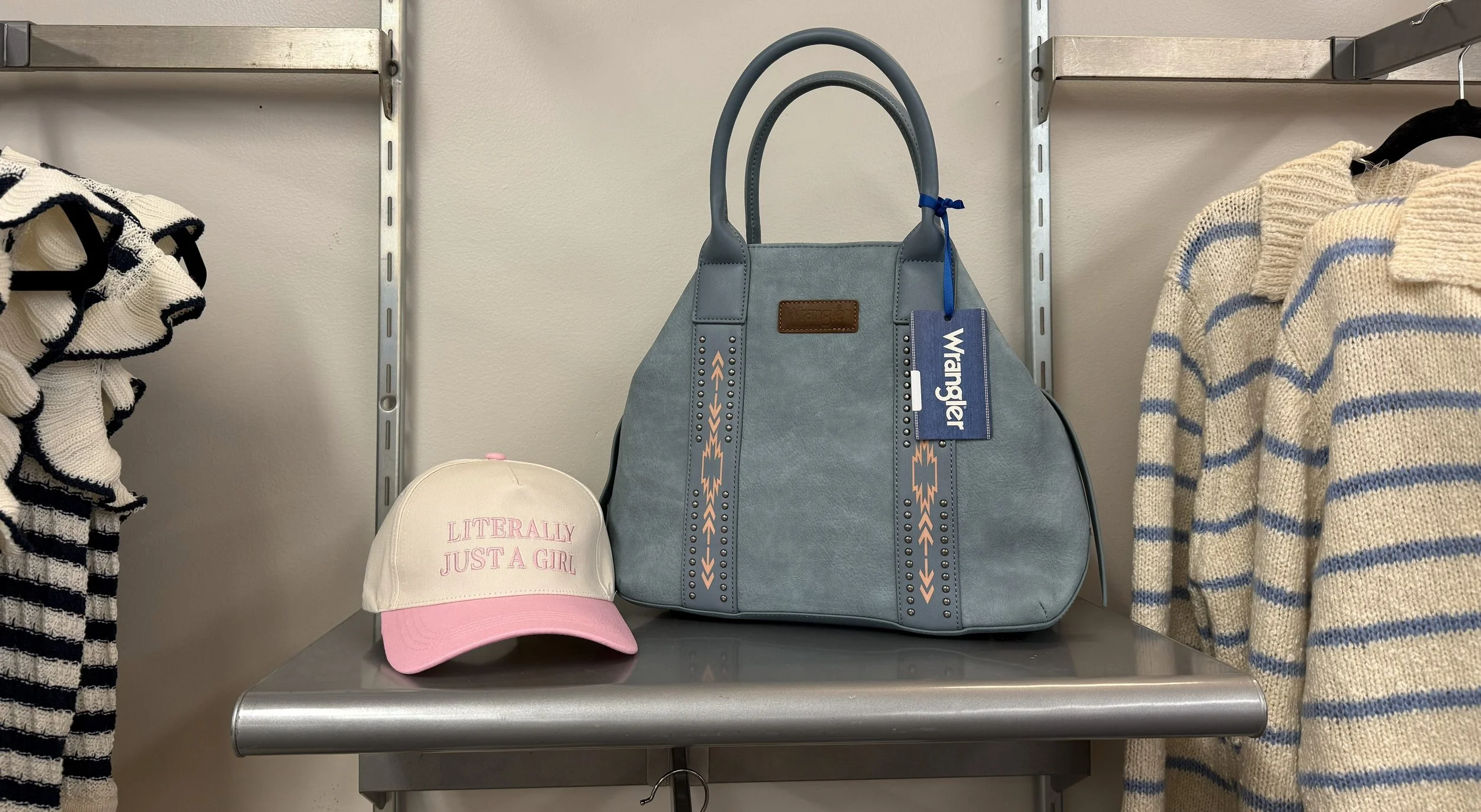 A blue handbag and a white and pink baseball cap with the text "LITERALLY JUST A GIRL" on it on a metal shelf in a store.