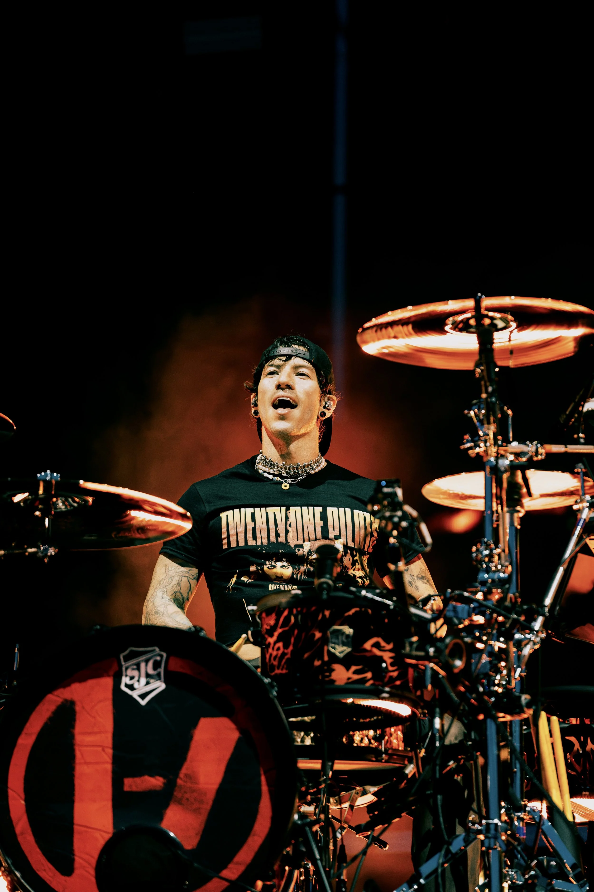 Joshua Dun from the band Twenty One Pilots performing at All Points East festival in Victoria Park,  wearing a black 'Twenty One Pilots' T-shirt, a black cap backwards, and layered necklaces, playing the drums on stage with red and orange lighting.