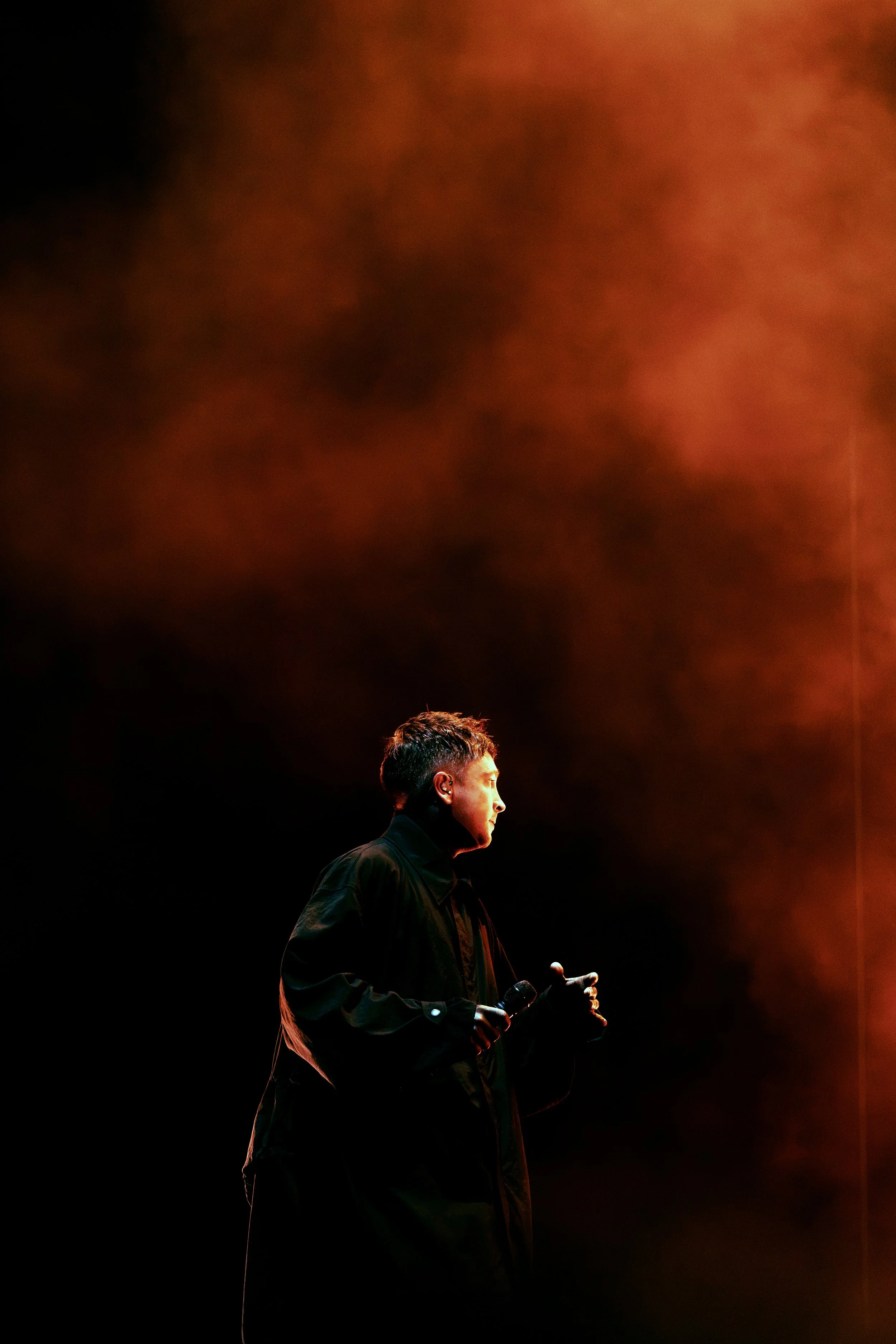 Tyler Joseph from the band Twenty One Pilots performing at All Points East festival in Victoria Park, London, 2026, holding a microphone stands on a dark stage illuminated by warm, orange stage lights above.