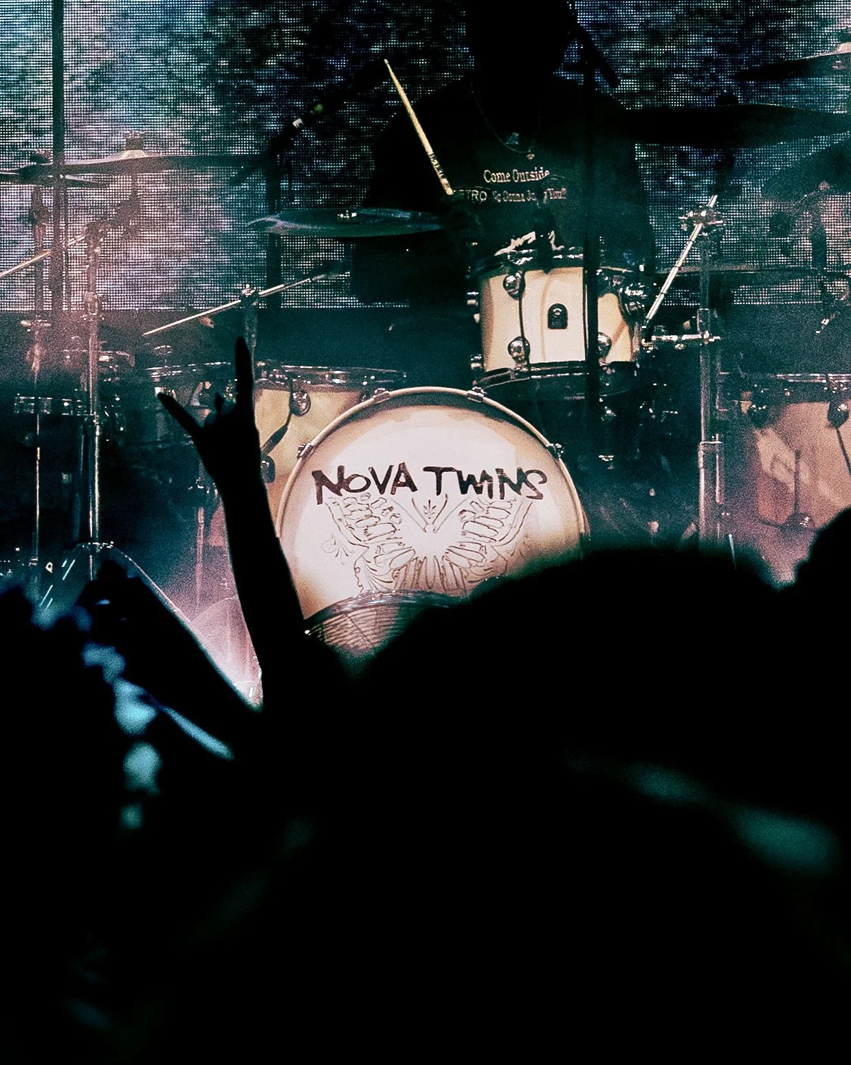 A person performing on stage in front of a drum set with the band's name, 'NOVA TWINS,' on the bass drum. The audience's silhouettes can be seen, with one person making a Rock and Roll hand gesture and holding a phone.
