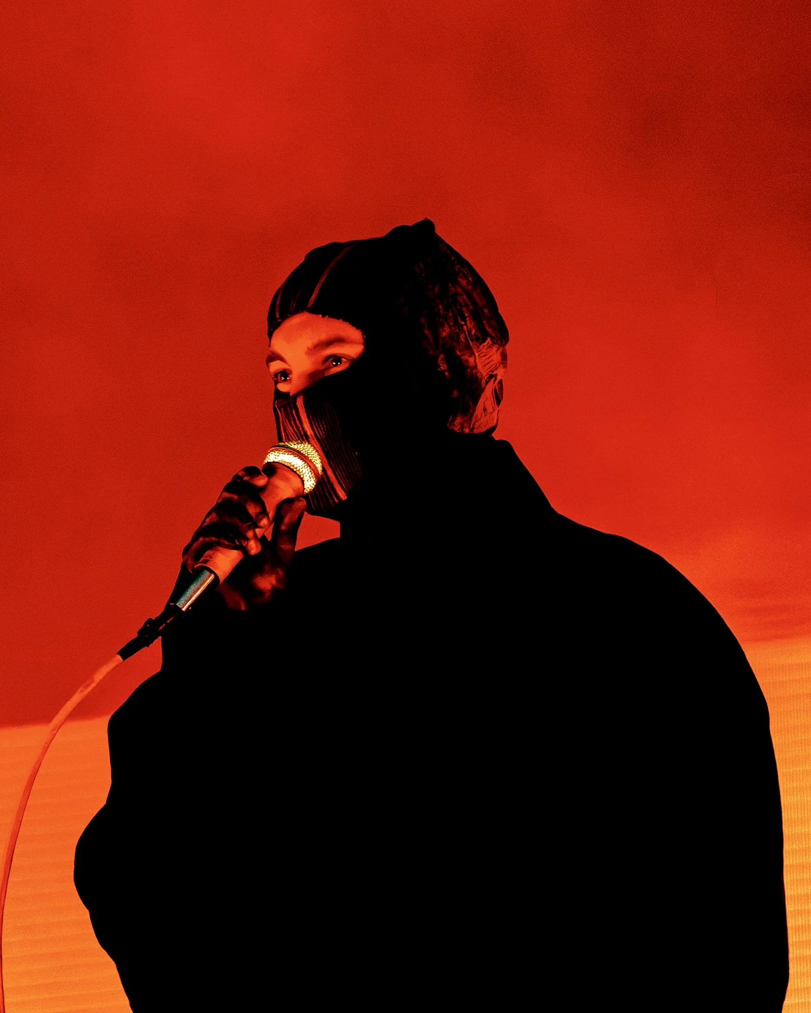 Tyler Joseph from the band Twenty One Pilots dressed in black with a hood covering most of their face, holding a microphone, against a red-orange background. Performing at All Points East festival in Victoria Park, London in 2026