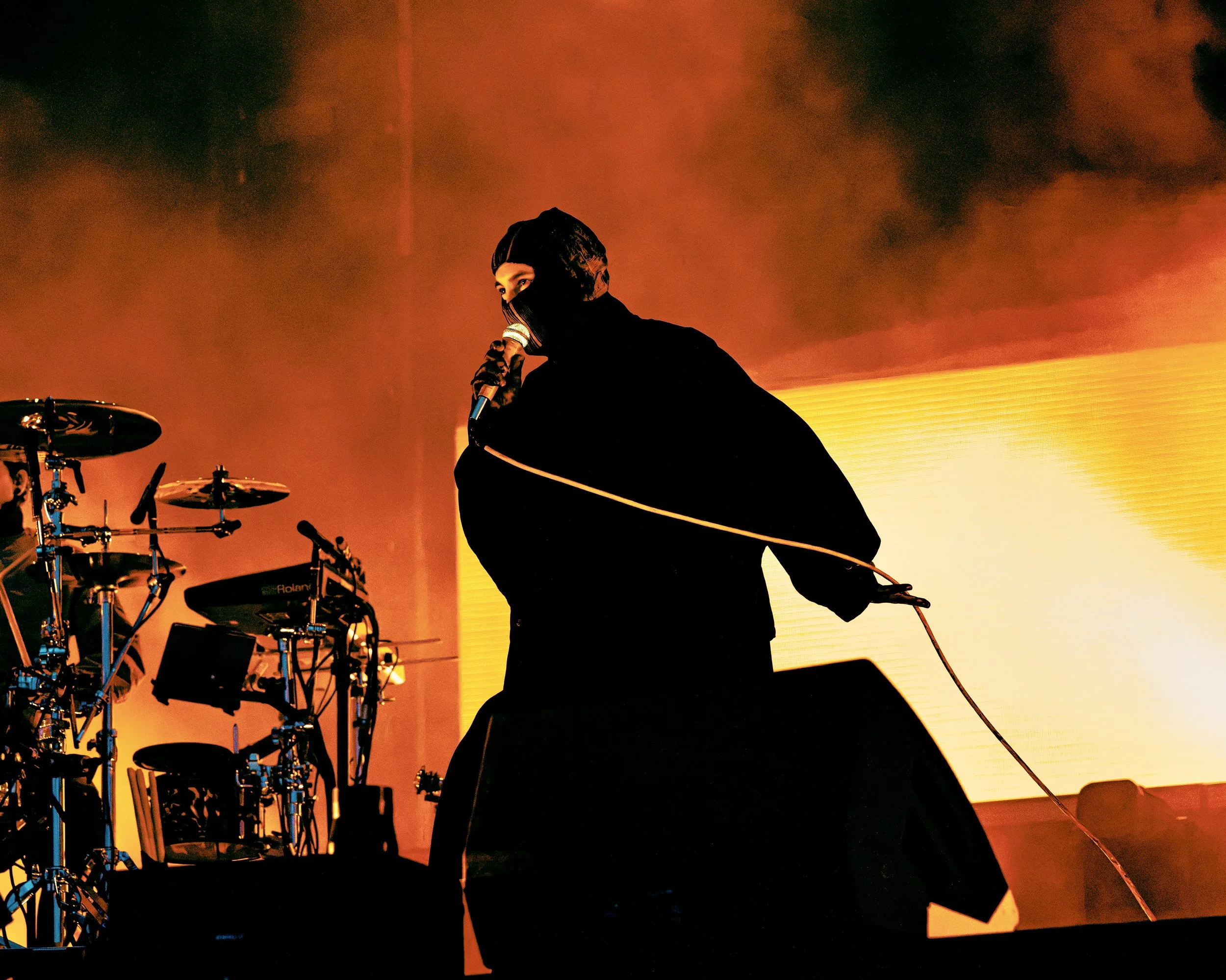 Tyler Jospeh from Twenty One Pilots performing at All Points East festival in Victoria Park, London, 2026 wearing a black hood and face mask, holding a microphone on stage with orange and yellow lighting, along with drum set in the background.