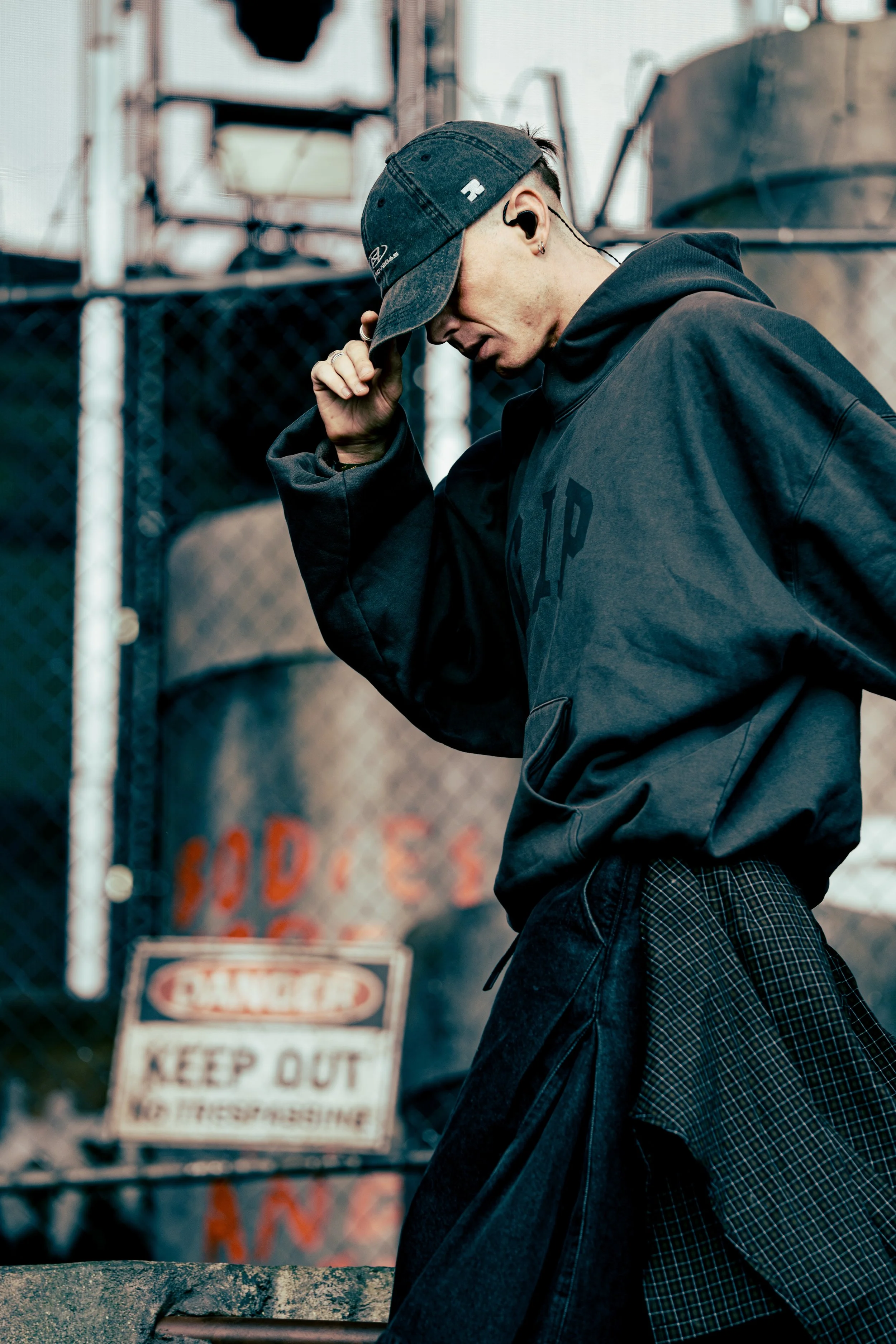 Musical artist Ren performing at All Points East festival in Victoria Park, London, 2026, wearing a dark hoodie, plaid shirt, and a baseball cap, adjusting the cap, standing near a chain-link fence with a blurred warning sign in the background.