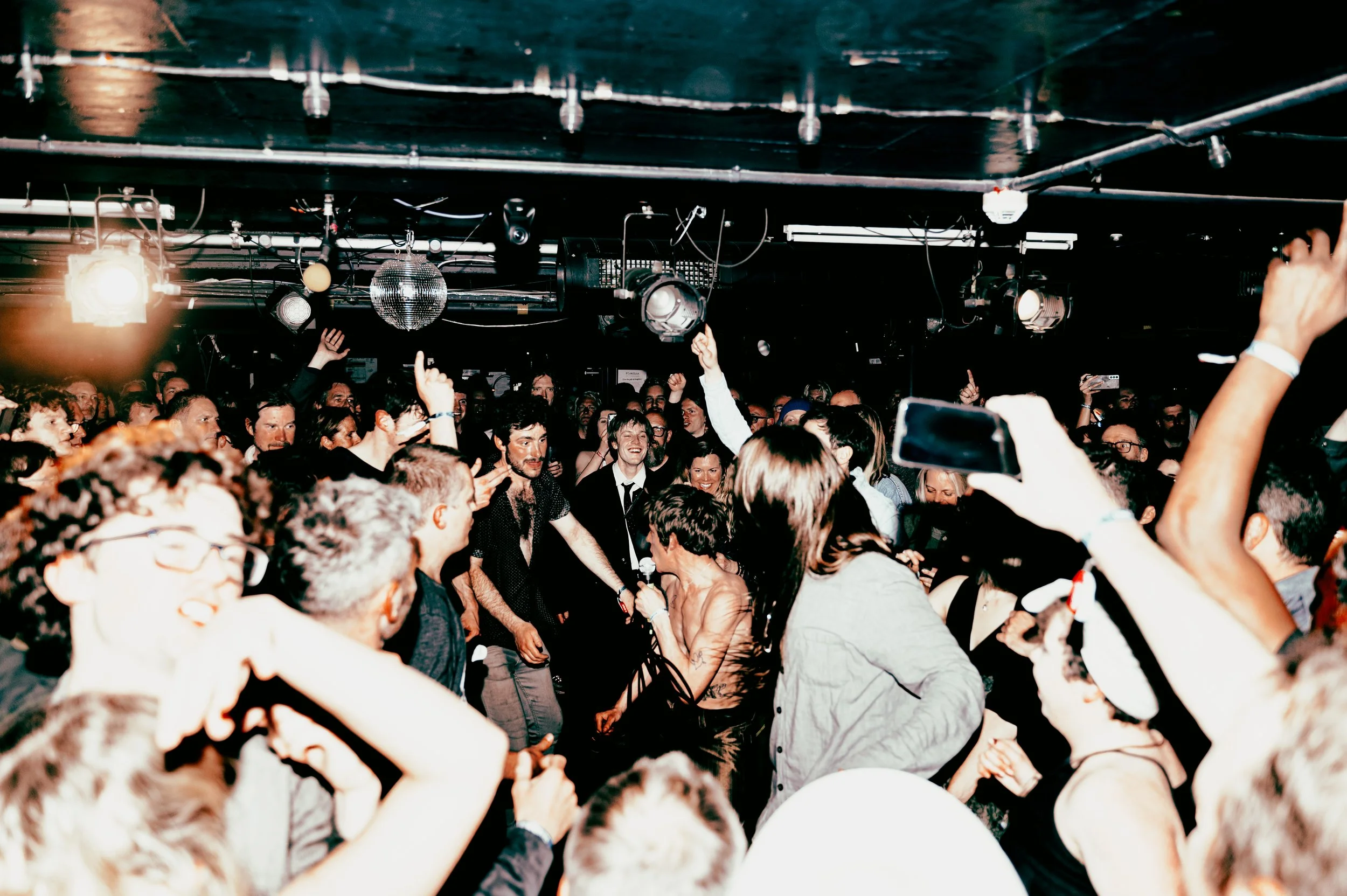A lively concert scene with a large crowd dancing and enjoying the music, stage lights, and a disco ball overhead. Zak, the frontman of the band Deadletter, is in the crowd in Komedia at The Great escape festival in Brighton.