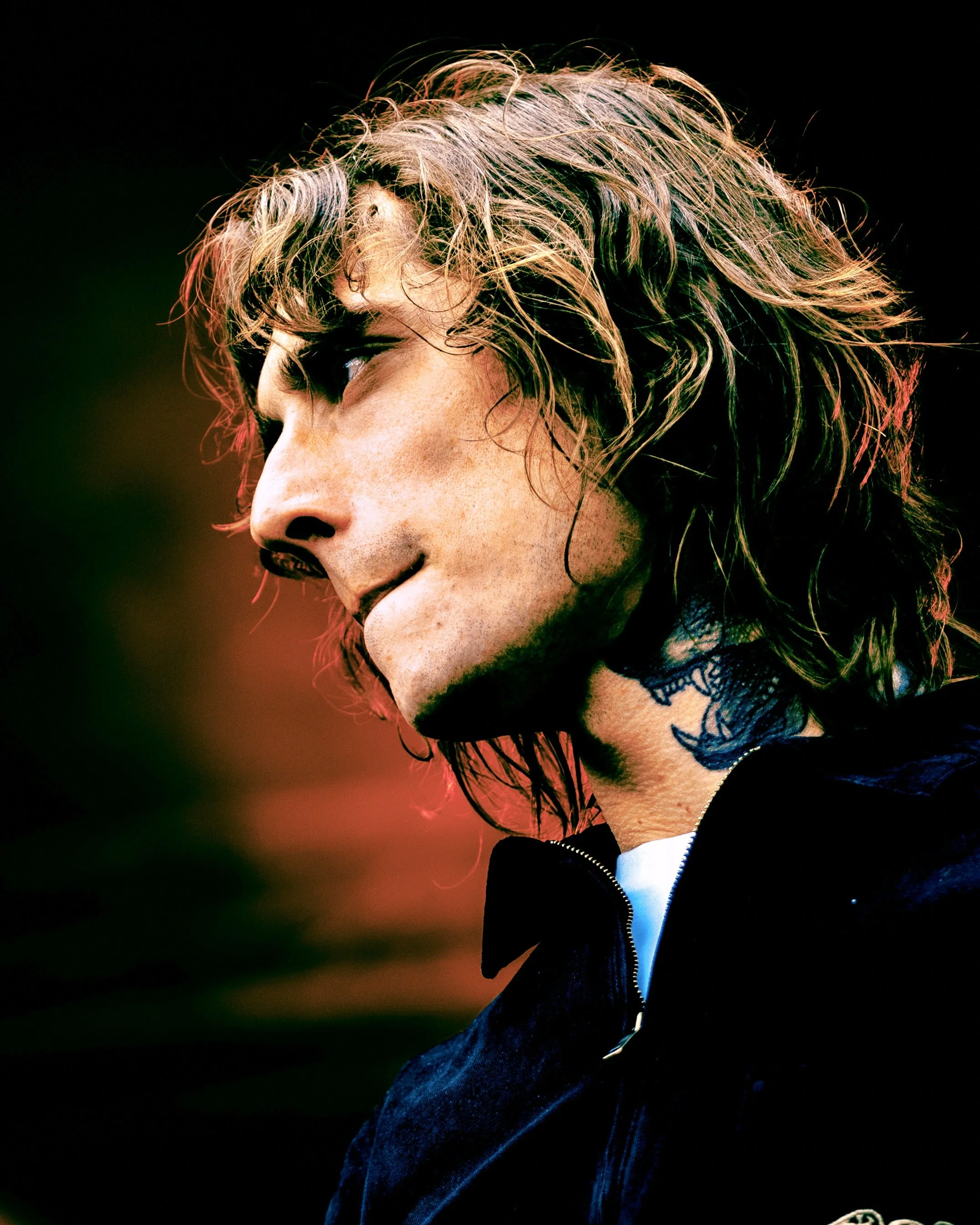 Close-up of a young man from the band Mouth Culture with curly hair, wearing a black jacket with a zipper, and tattoos visible on his neck, set against a dark background. He is on stage at All Points East festival in Victoria Park, London in 2026