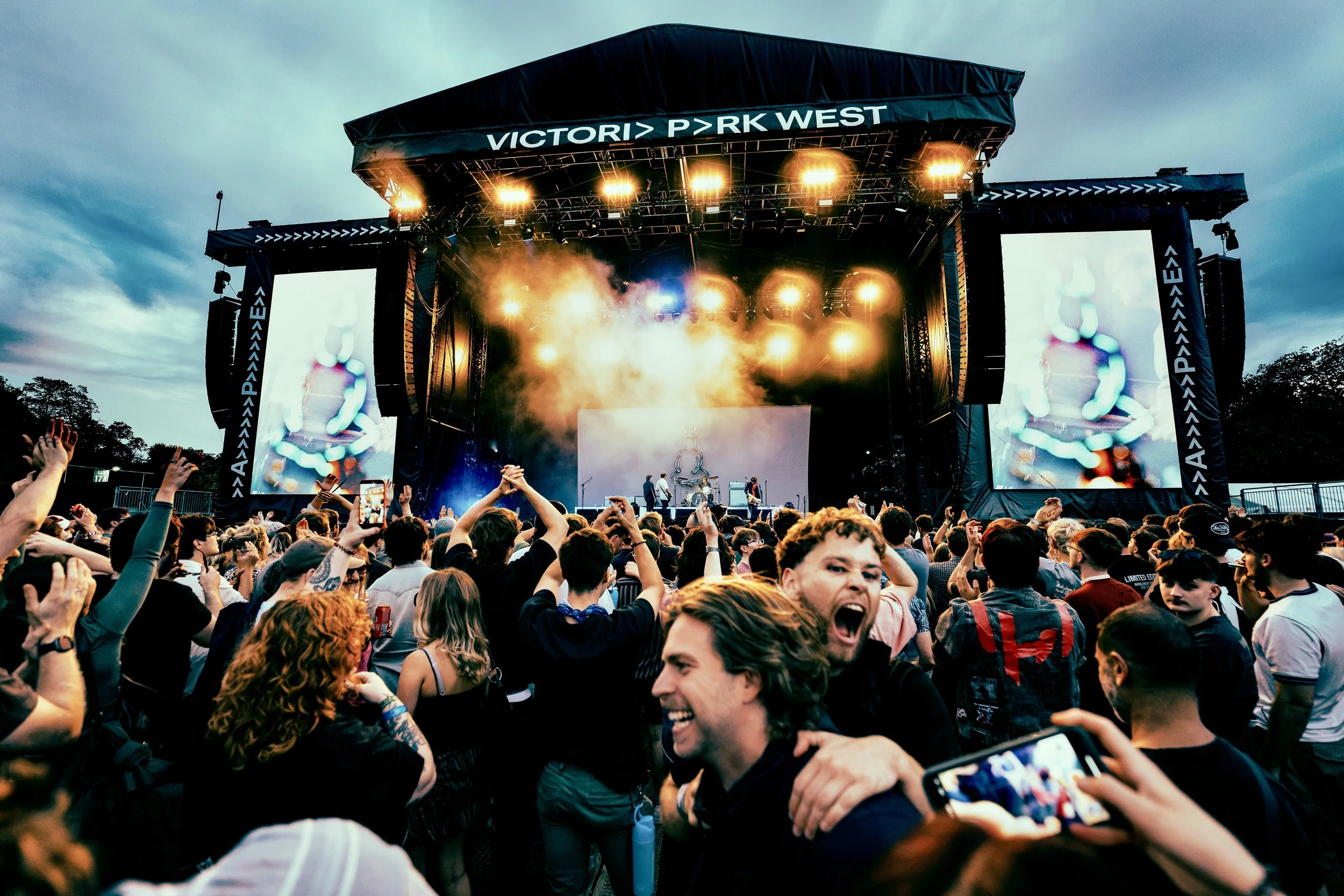 A large crowd of people attending All Points East festival in Victoria Park, London in 2026. The band Wunderhorse is on stage, featuring bright lights and screens, and an overcast sky in the background.