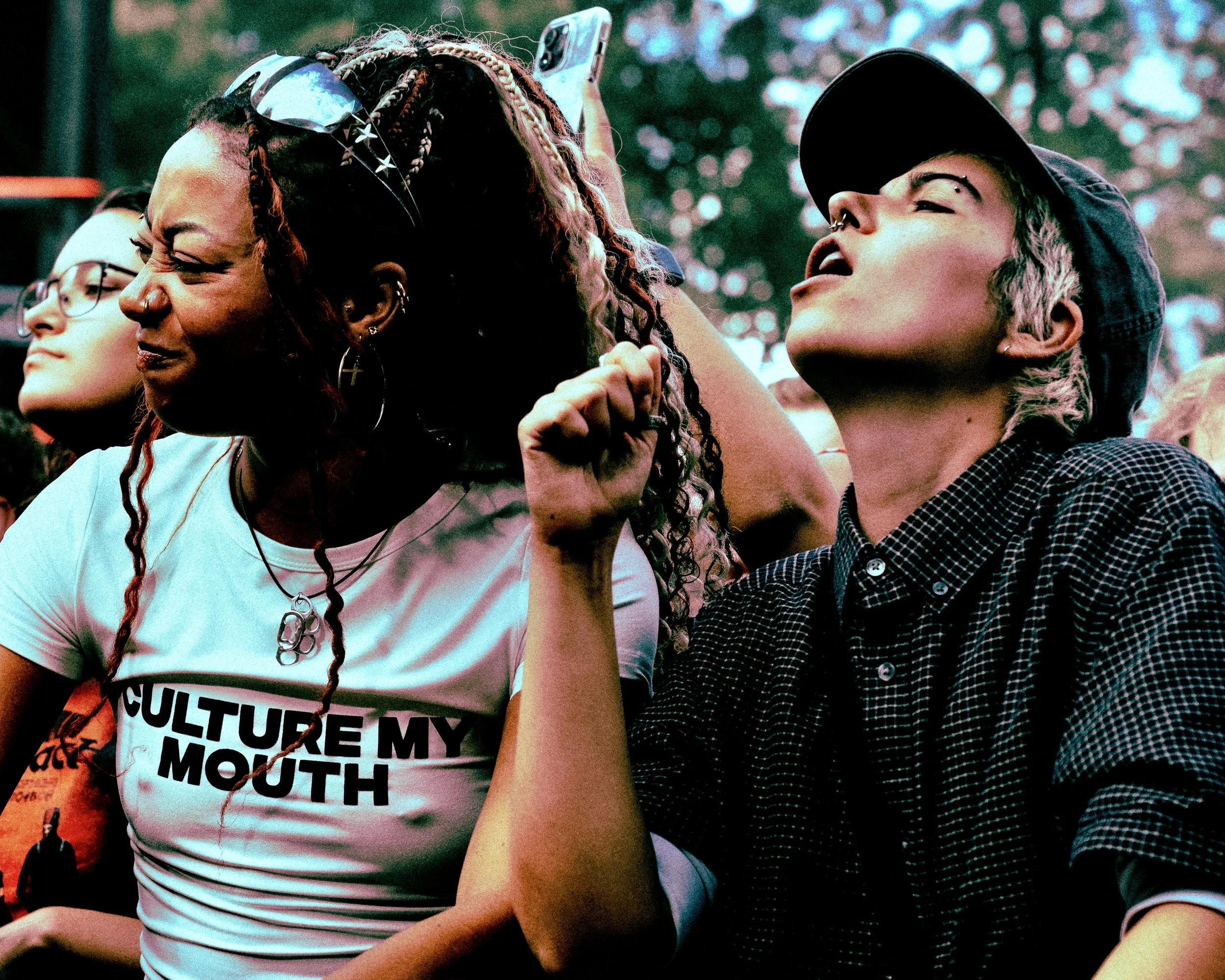 Close-up of two people at the barricade at a music festival, one with braided hair and sunglasses, holding a smartphone, and the other with a cap and piercings, outdoors with blurred trees in the background. They are in the audience of Mouth Culture