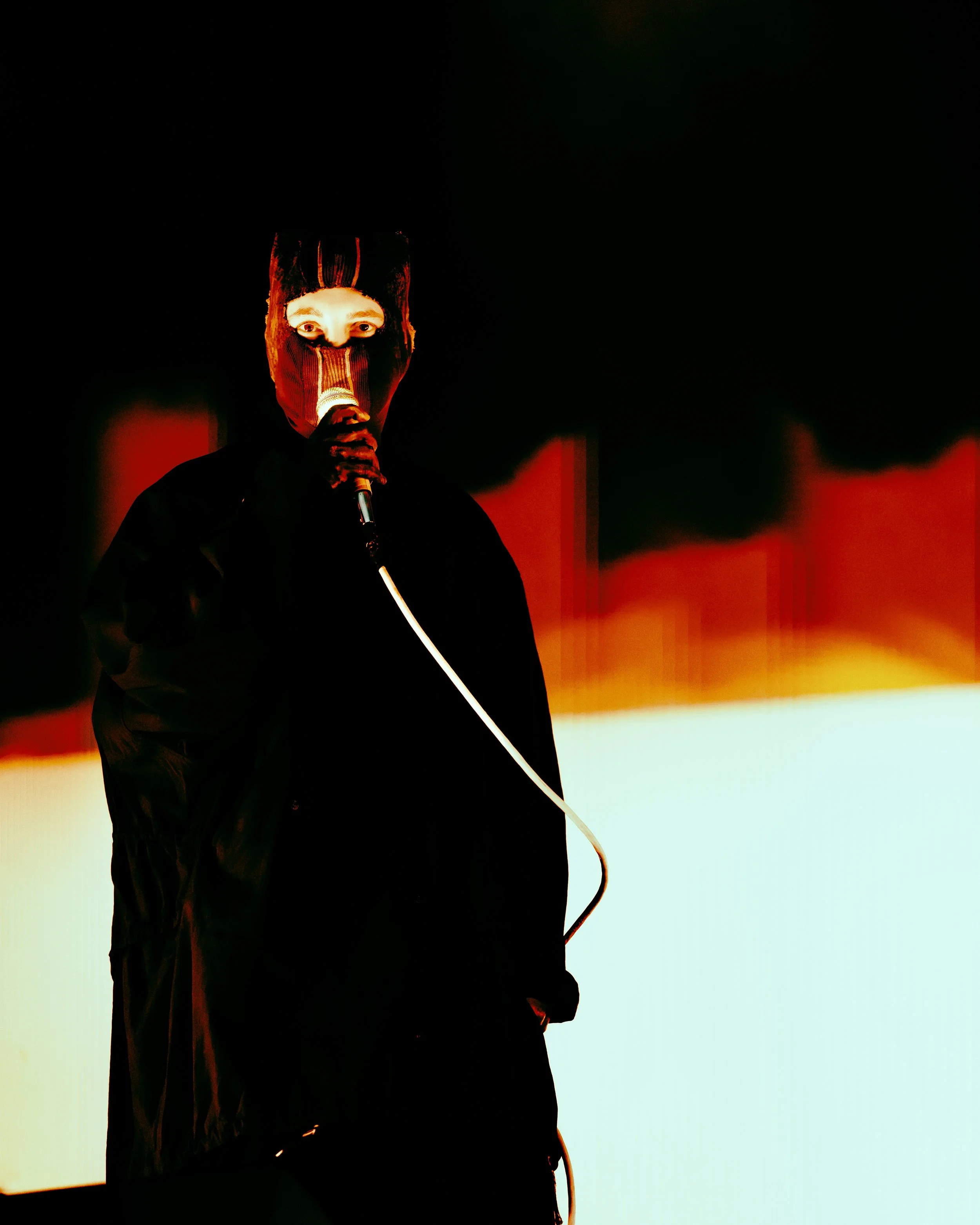 Tyler Joseph from the band Twenty One Pilots performing at All Points East festival in Victoria Park, London in 2026, wearing a ski mask holding a microphone, standing in front of a colorful, abstract background with black, red, and yellow hues.