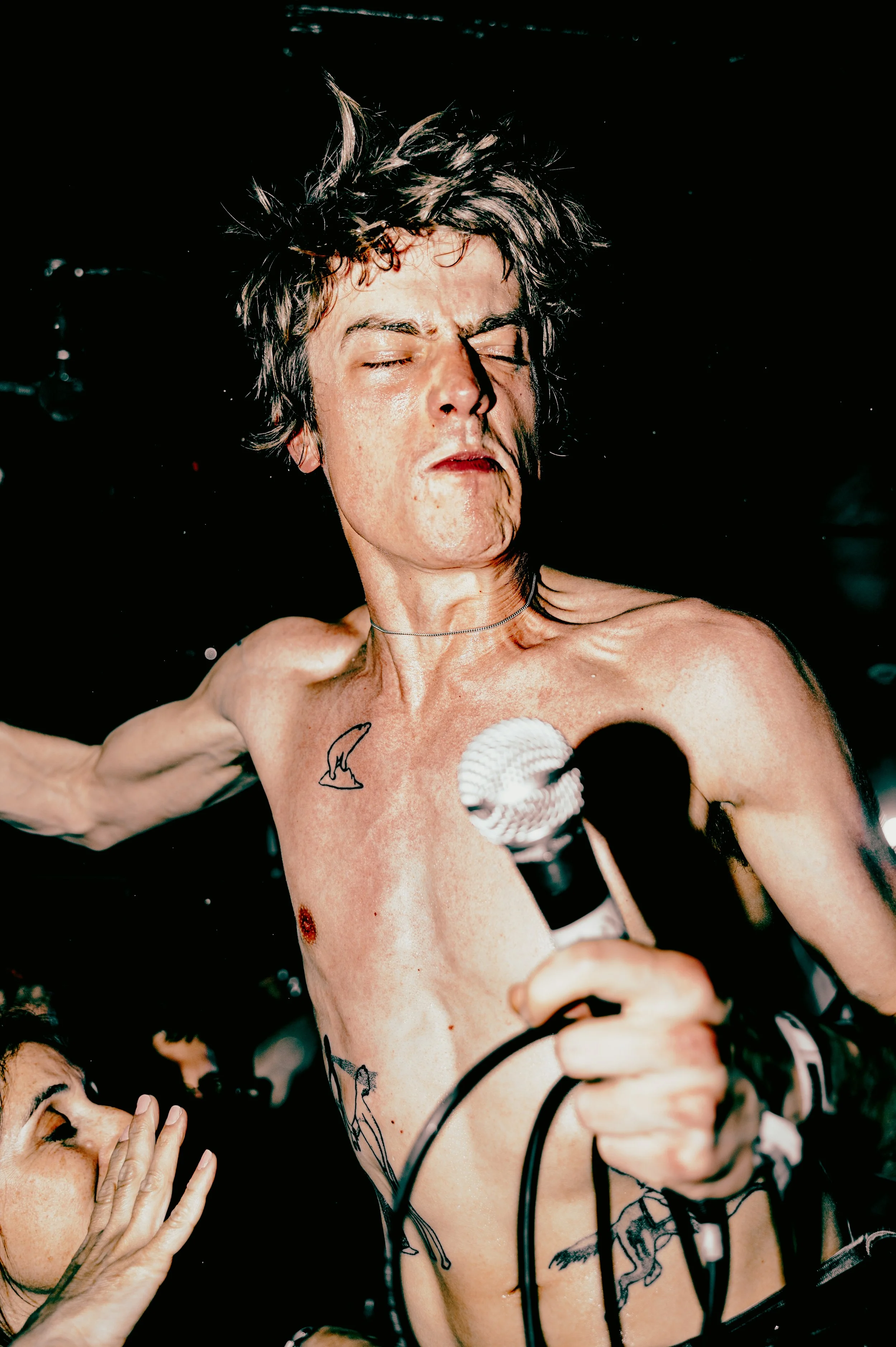 A shirtless person with wet hair and tattoos, holding a microphone and singing passionately, surrounded by fans touching their shoulder. The man is Zak, frontman of the band Deadletter performing at Komedia in Brighton during The Great Escape Festiva