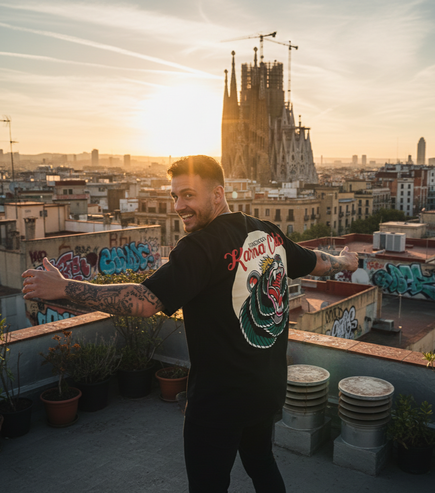 A young man with tattoos smiling and extending his arms on a rooftop in Barcelona during sunset, with the Sagrada Família in the background and graffiti on the surrounding walls.