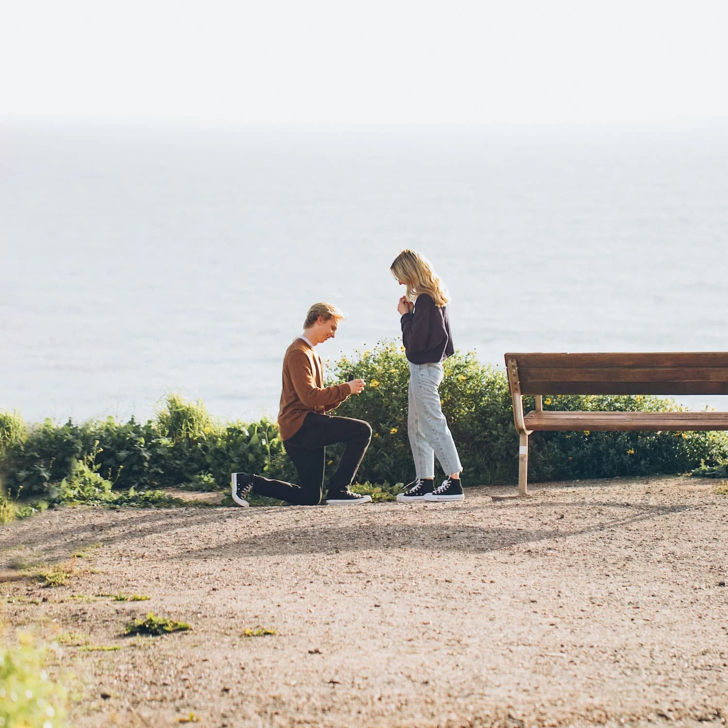 Boyfriend proposing to girlfriend by the ocean in a budget friendly idea, kneeling with engagement ring