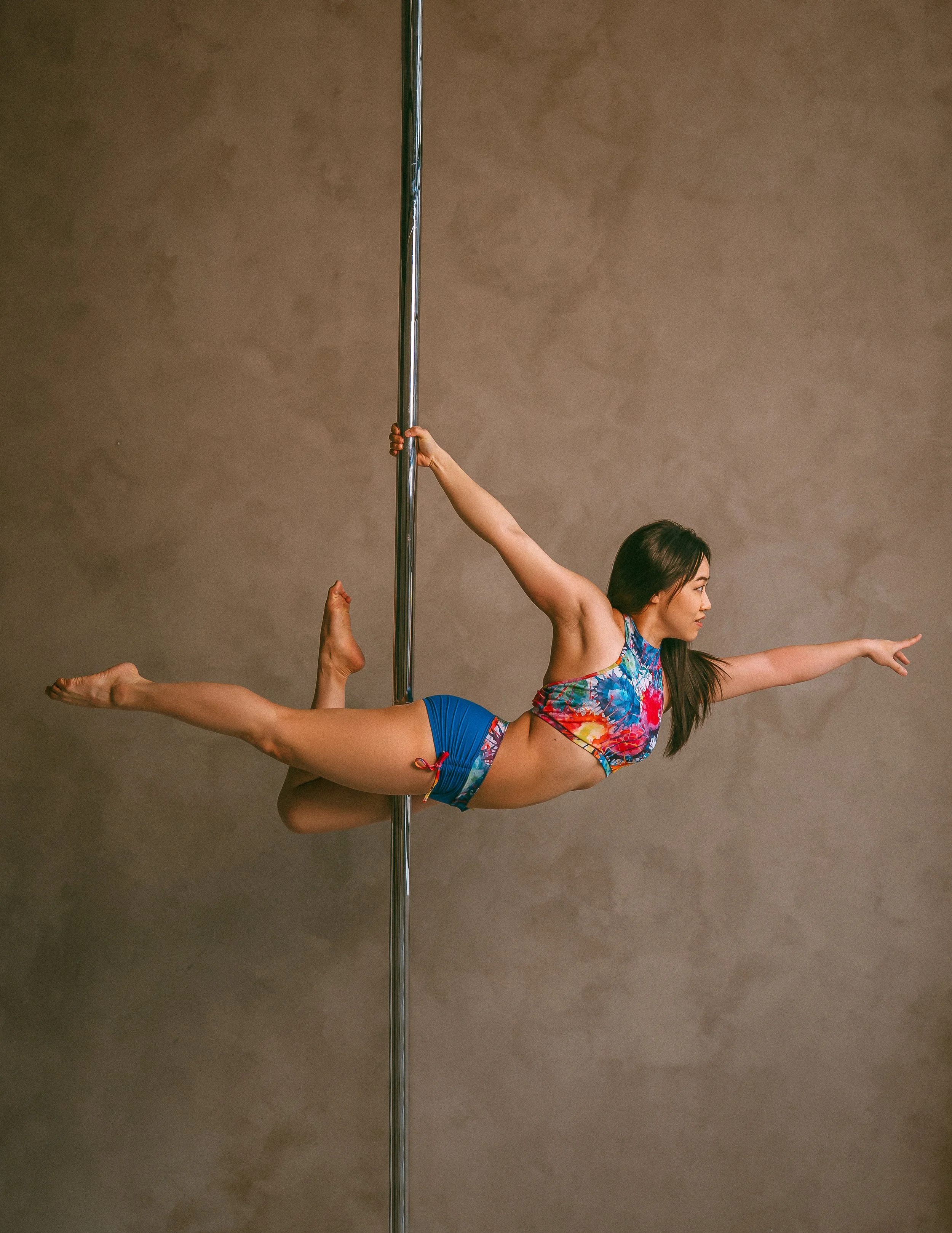 A woman performing a pole dance move, hanging horizontally with her arms and legs extended, holding onto a vertical dance pole against a plain gray background.