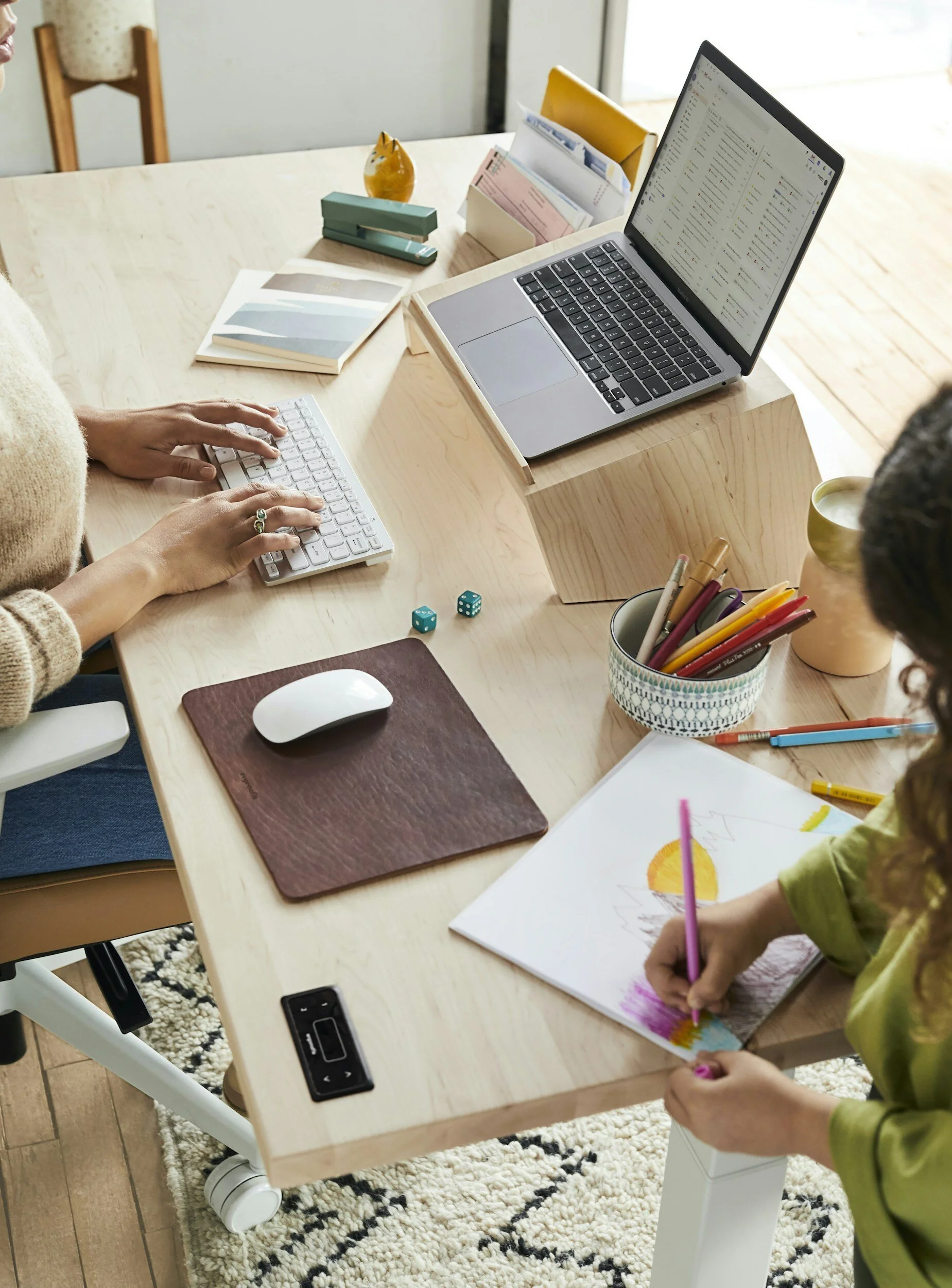 A workspace with two people, one typing on a keyboard, the other drawing with colored pencils. The desk has a laptop, a mouse, some notebooks, and art supplies. Two dice are on the desk, and various stationery items are organized in containers.