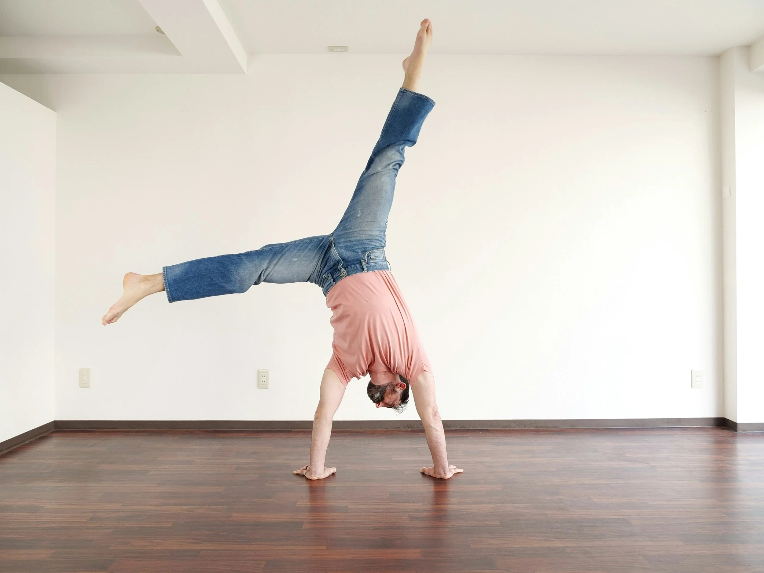 A man performing a handstand in an empty room with wooden floors, wearing a pink shirt and blue jeans.