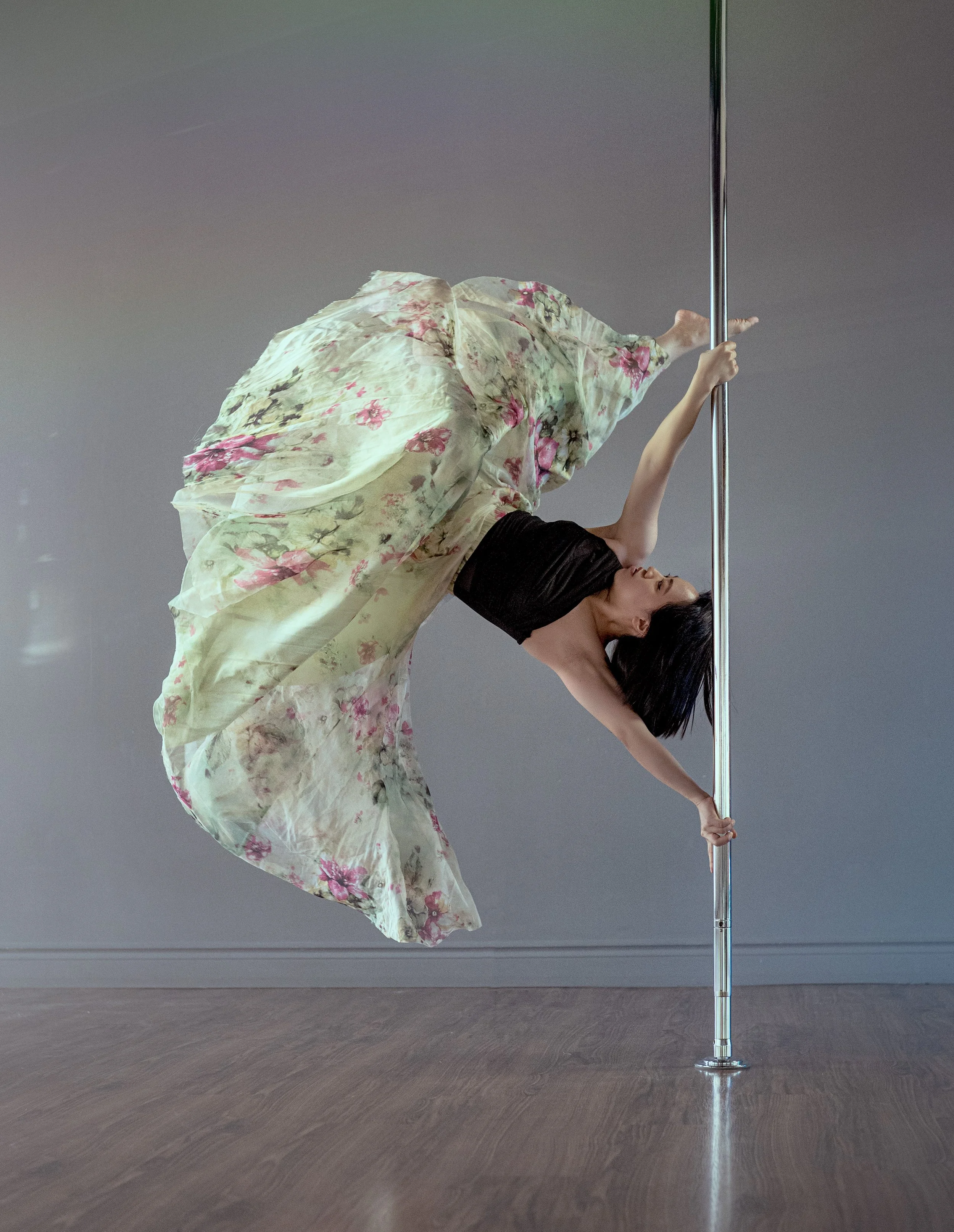 A woman performing a pole dance move, bent backward holding the pole with one hand and foot, dressed in a black top and a flowing floral skirt, in a minimalist room with a gray wall and wooden floor.