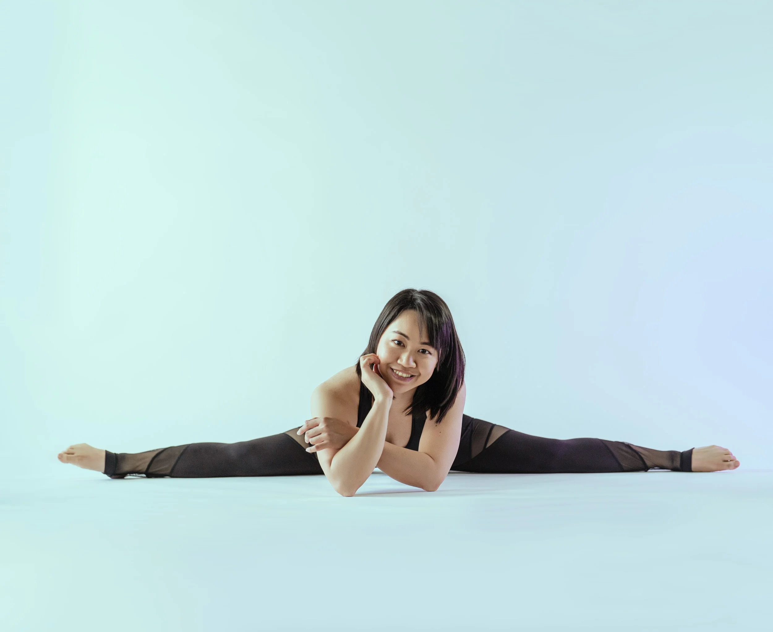 Young woman in black athletic wear performing full split on the floor, smiling, against a light background.