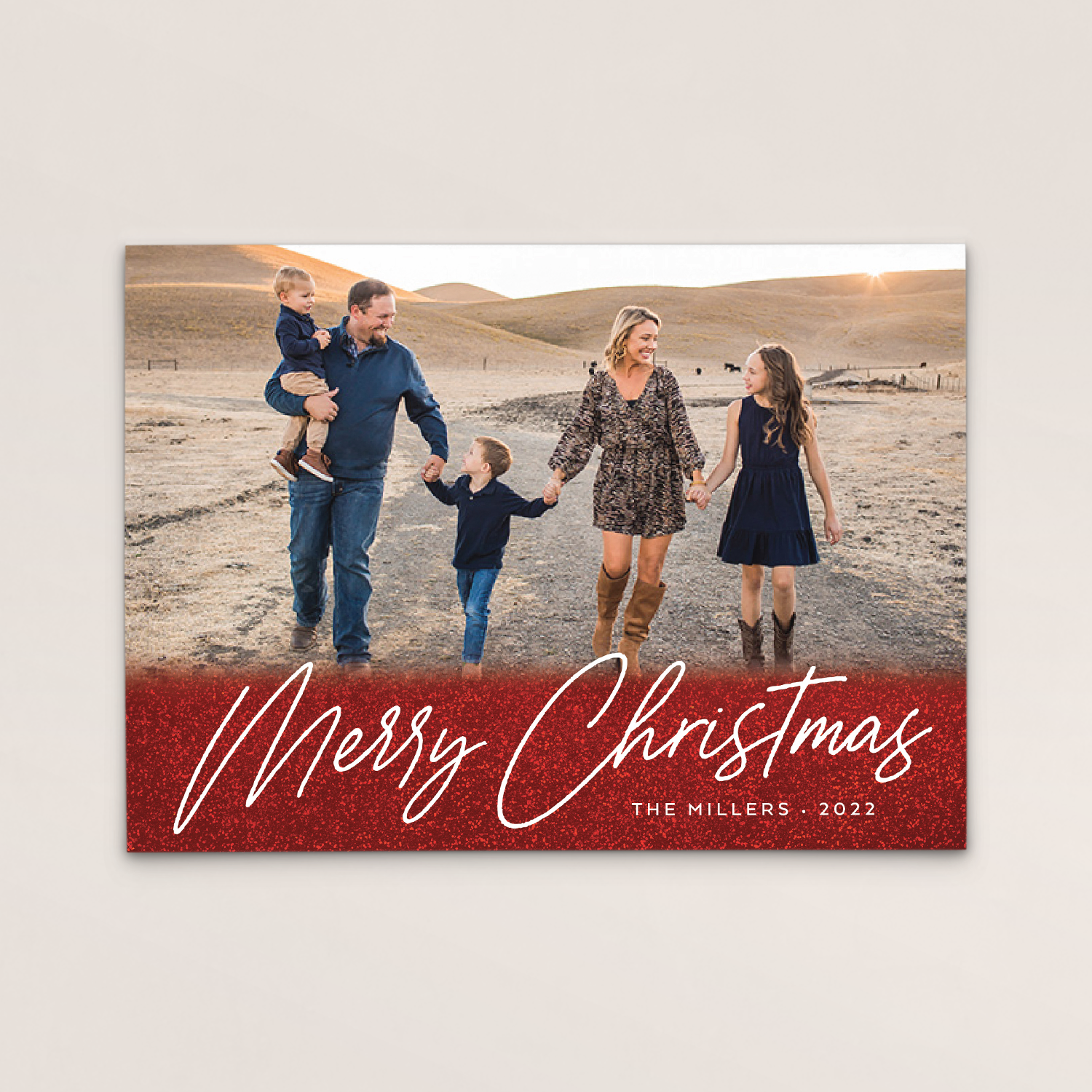 Family walking outdoors in a rural landscape during sunset, holding hands and smiling, with a red banner at the bottom reading "Merry Christmas" and text indicating the family name "The Millers" and year "2022."