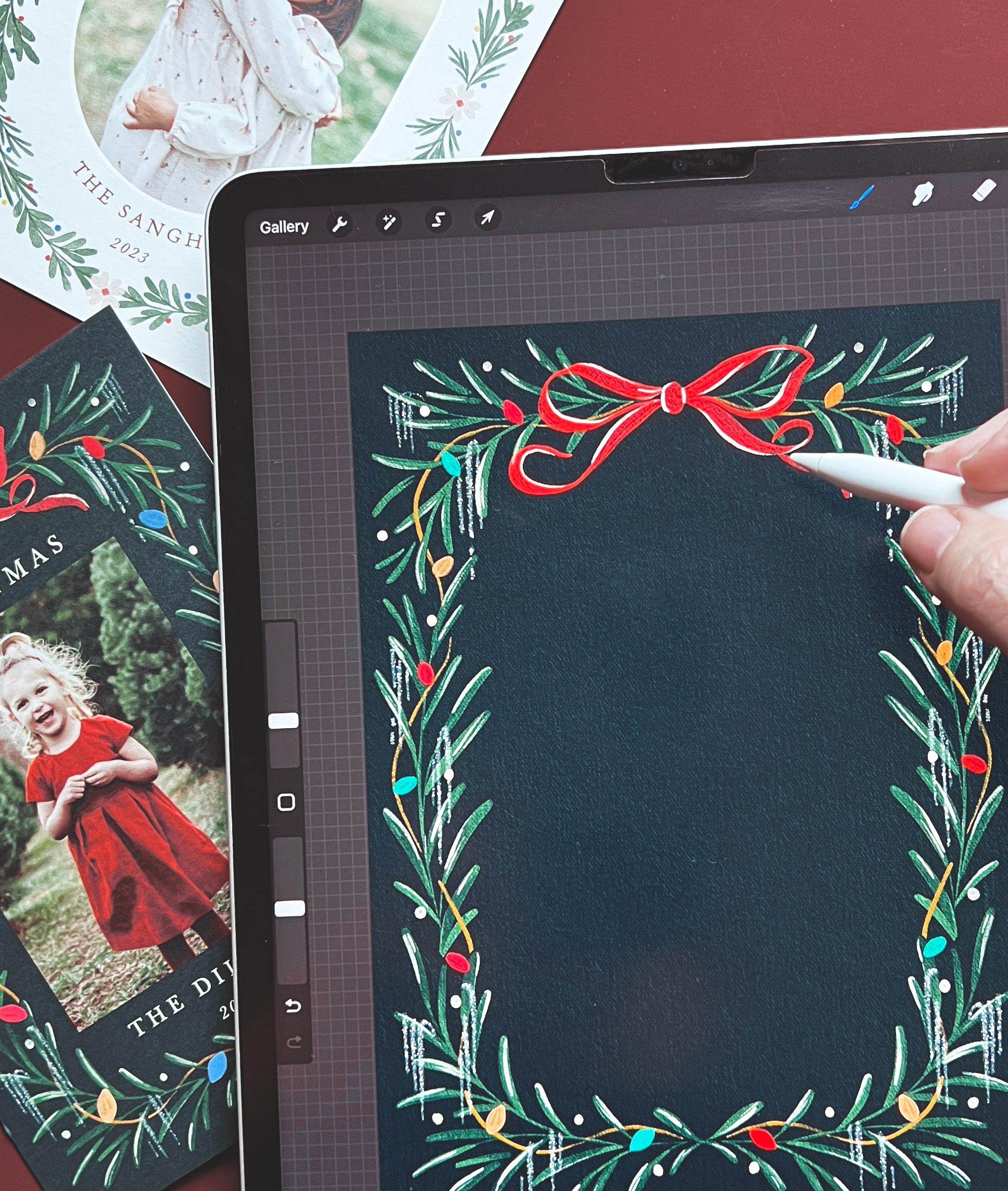 Digital device screen displaying a holiday-themed illustration of a green pine branch decorated with multi-colored lights, red bow, and white accents, being drawn with a stylus. Surrounding papers include a holiday card with green foliage border and a partial photo of a girl in a red dress.