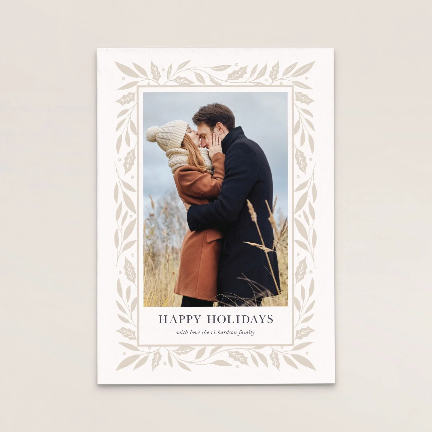Holiday greeting card with a photo of a couple embracing outdoors, surrounded by tall grass and bushes, with text that reads 'HAPPY HOLIDAYS with love the richardson family' and a decorative leaf border.