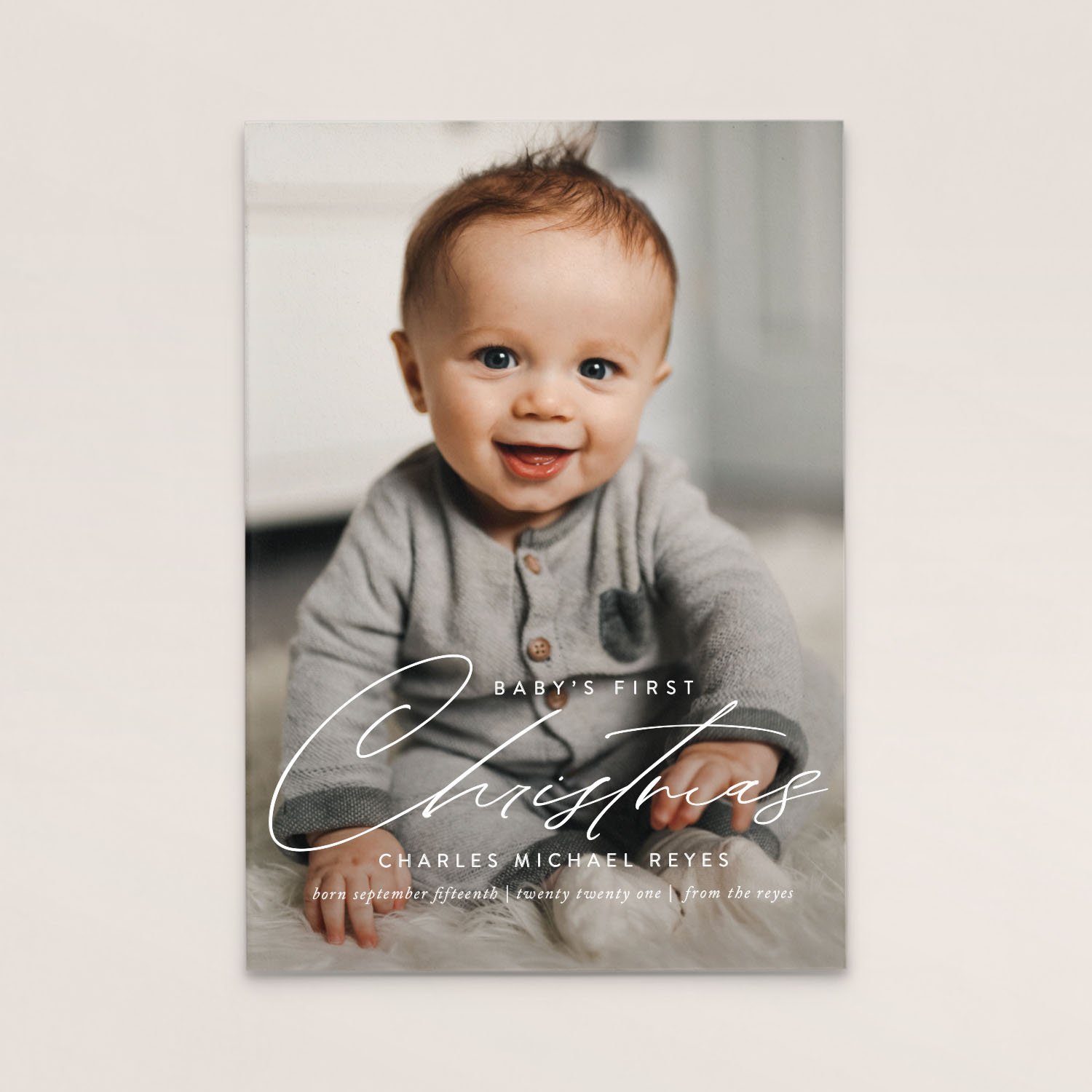 A Christmas card featuring a smiling baby with blue eyes, wearing a gray onesie, sitting on a soft white surface. The card reads 'Baby's First Christmas' and includes the name Charles Michael Reyes, born September 15th, 2021.