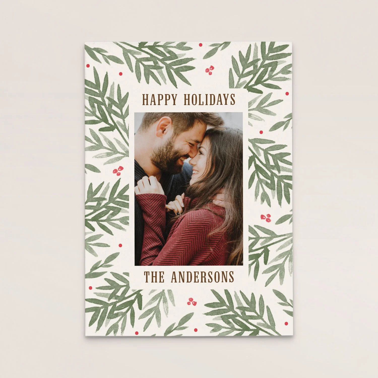 Holiday greeting card with a photo of a smiling couple touching foreheads, surrounded by green leaves and red berries, and the text 'Happy Holidays' and 'The Andersons'.