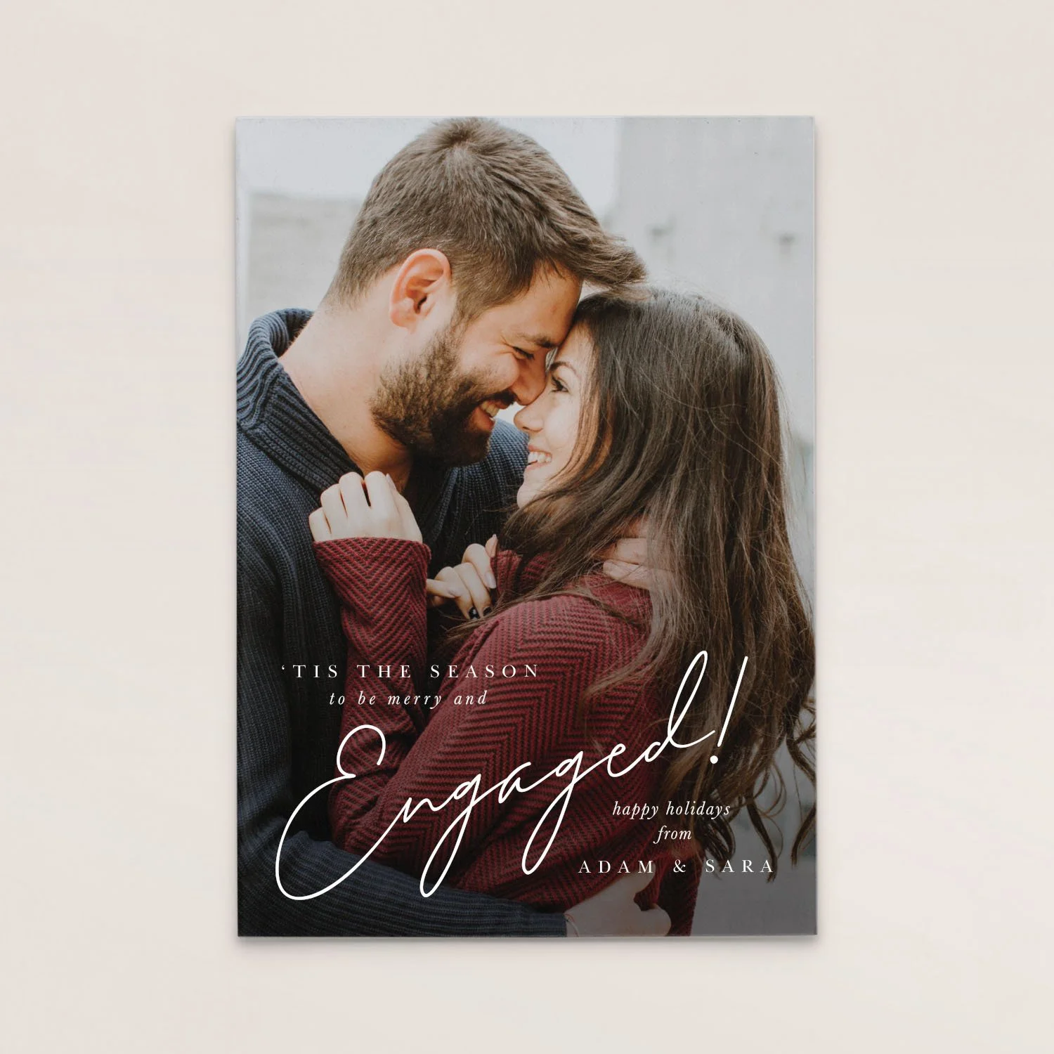 Holiday greeting card with a photo of a couple smiling and touching foreheads, dressed in winter clothing, with the text 'Tis the season to be merry and engaged! Happy holidays from Adam & Sara'.