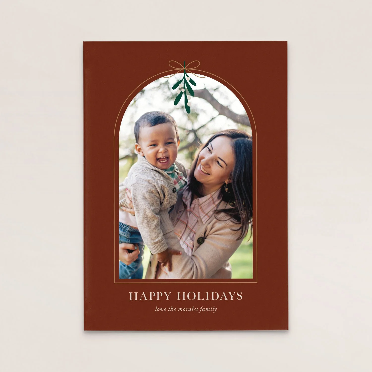 Holiday greeting card with a photo of a woman holding a smiling child outdoors, framed by a decorative border with botanical elements, and the text 'HAPPY HOLIDAYS' and 'love the morales family' at the bottom.
