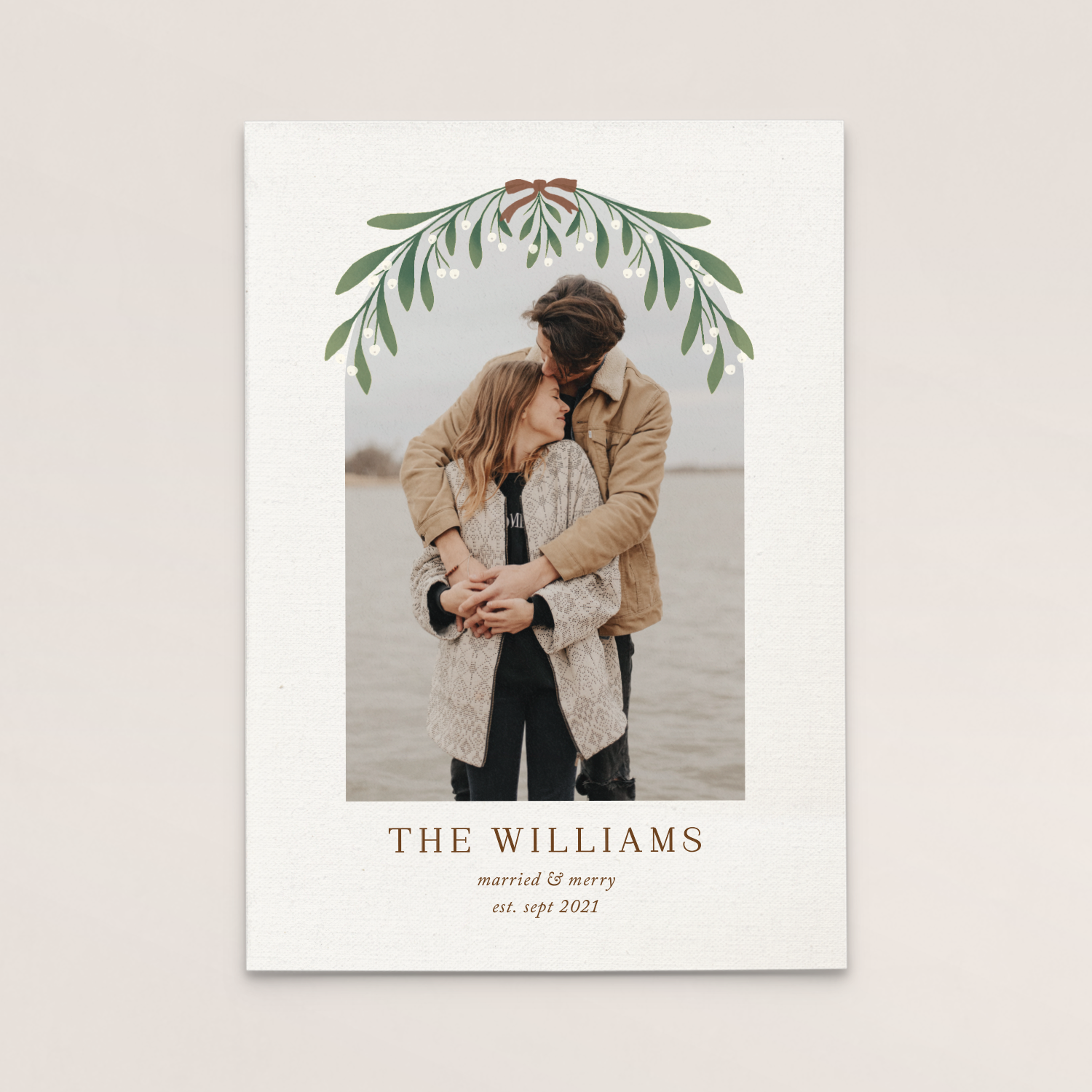 A wedding announcement card featuring a romantic photo of a couple embracing and kissing outdoors at the beach, with a green floral border at the top and text below stating the couple's last name, wedding details, and date.