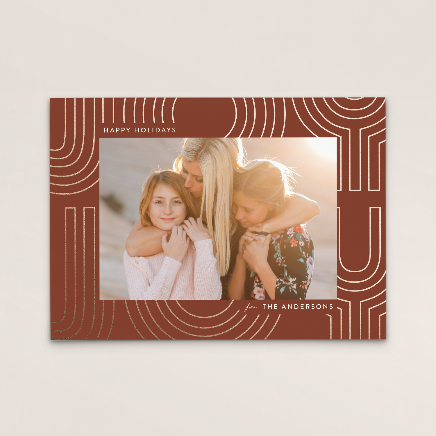 Holiday greeting card with a photo of a woman and two young girls hugging, surrounded by a decorative brown border with white lines, and the text "HAPPY HOLIDAYS" and "from THE ANDERSONS".