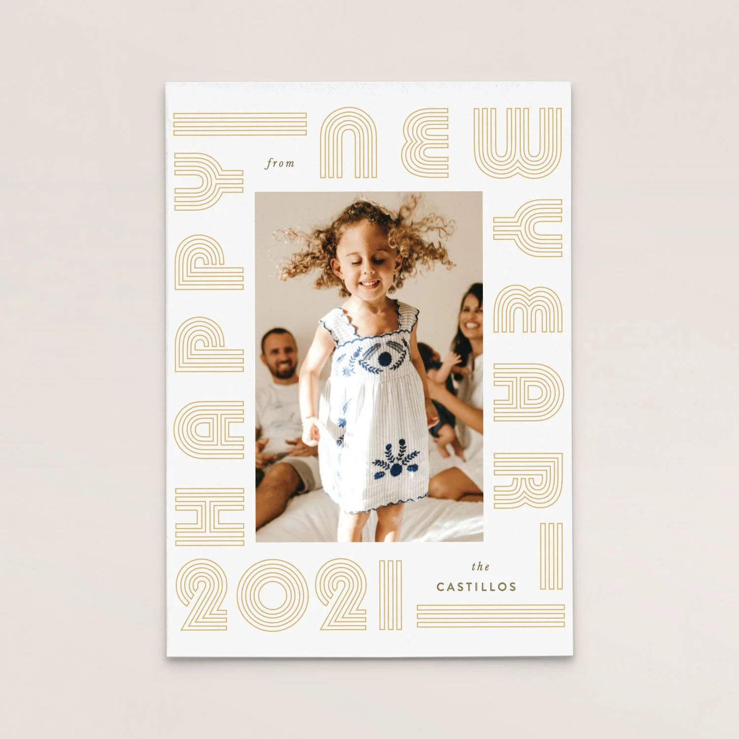 A family photo with a young girl wearing a white dress with blue embroidery, standing on a bed, with smiling adults in the background. The picture is framed by a white border with gold text reading "PRESENTED FROM THE CASTILLOS 2021" and other decora