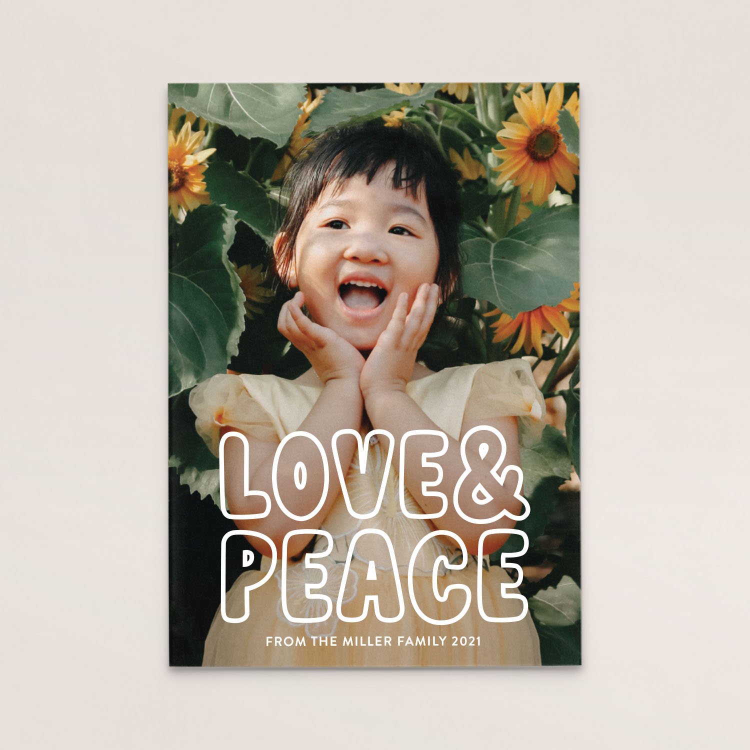 A young girl with black hair, wearing a beige dress, holding her face with her hands and smiling, in front of sunflower plants. The text on the image reads 'LOVE & PEACE' and 'FROM THE MILLER FAMILY 2021'.
