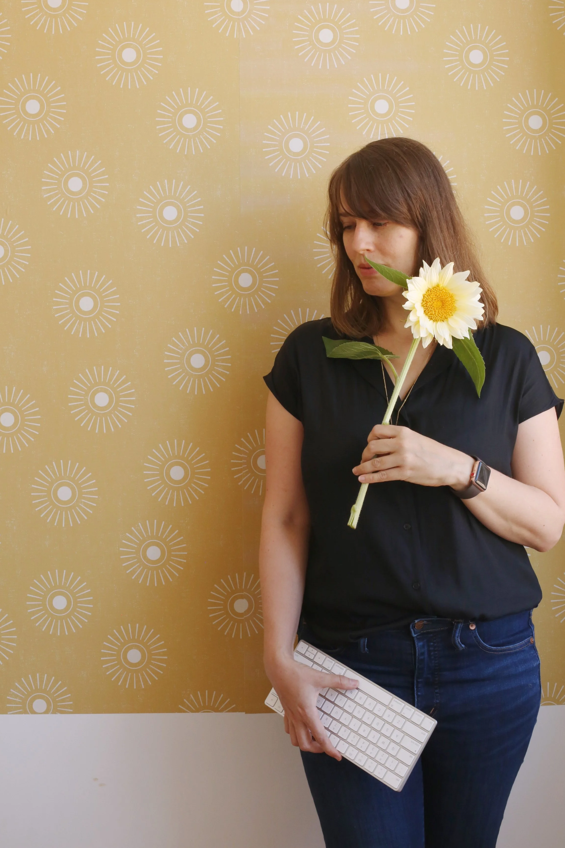 A woman with brown hair wearing a black shirt and jeans, holding a white computer keyboard, standing against a yellow wall with a white circular pattern. She is holding a daisy flower near her face.