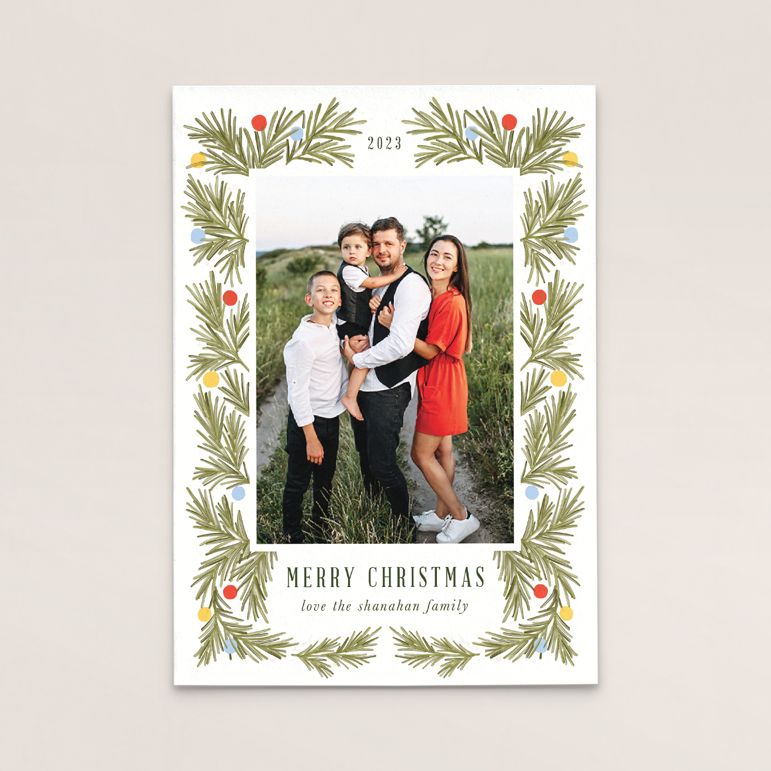 A Christmas greeting card with a family photo showing a woman, man, and two boys standing outdoors in a field, surrounded by a decorative border of pine branches and colored dots, with text that reads, 'Merry Christmas,' 'love the shanahan family,' a