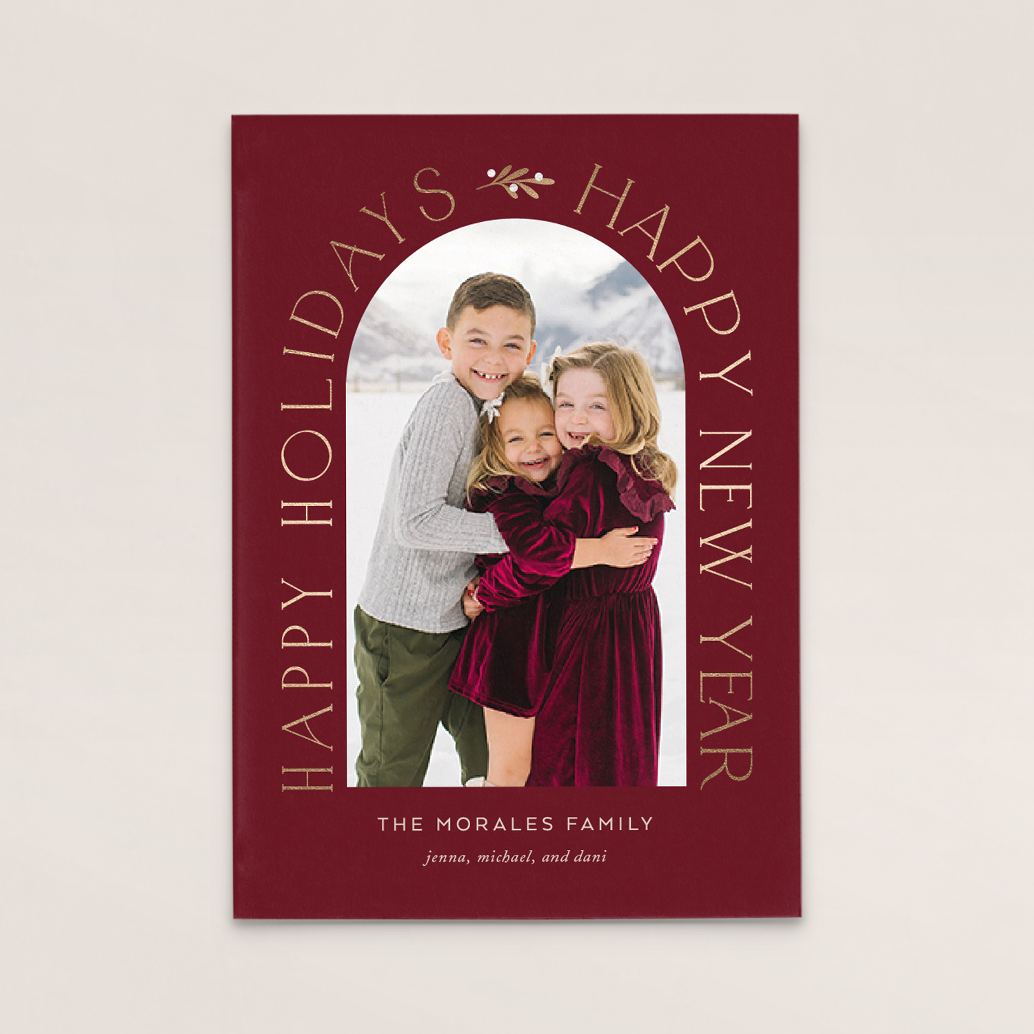 Family photo of three children smiling outdoors in snowy mountains, framed by a red border with gold text reading 'Happy Holidays' and 'The Morales Family' with their names.