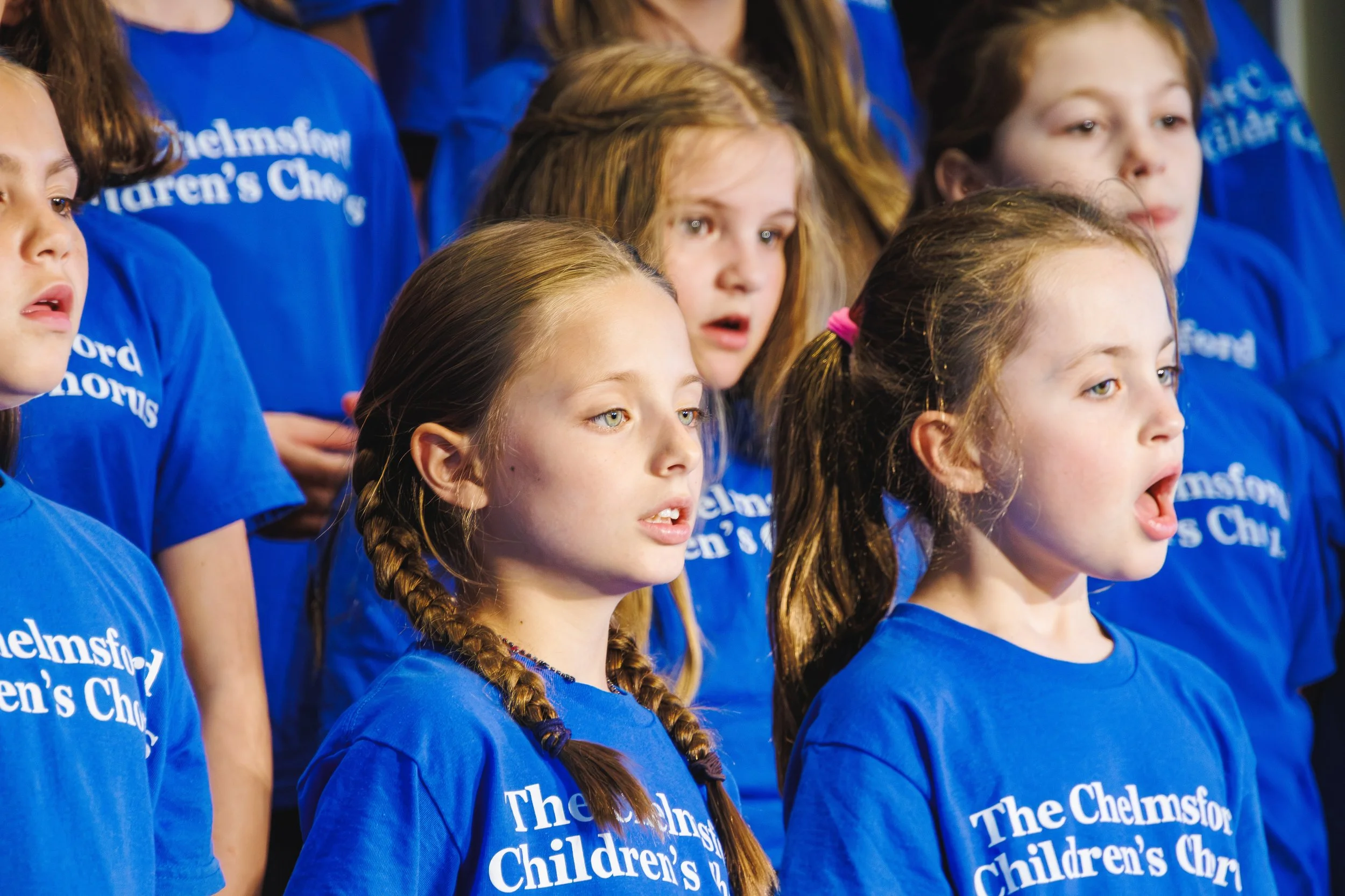 A group of young girls performing on stage during a choir recital, dressed in black, with various expressions of excitement and anticipation.
