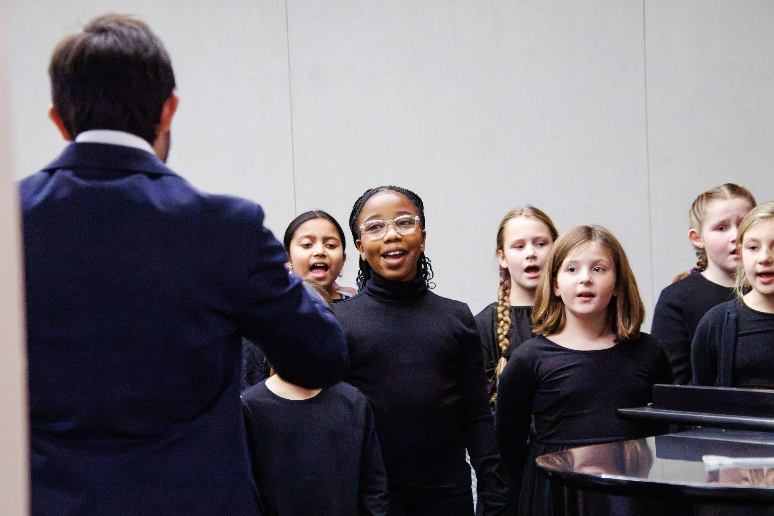 A group of young girls performing on stage during a choir recital, dressed in black, with various expressions of excitement and anticipation.