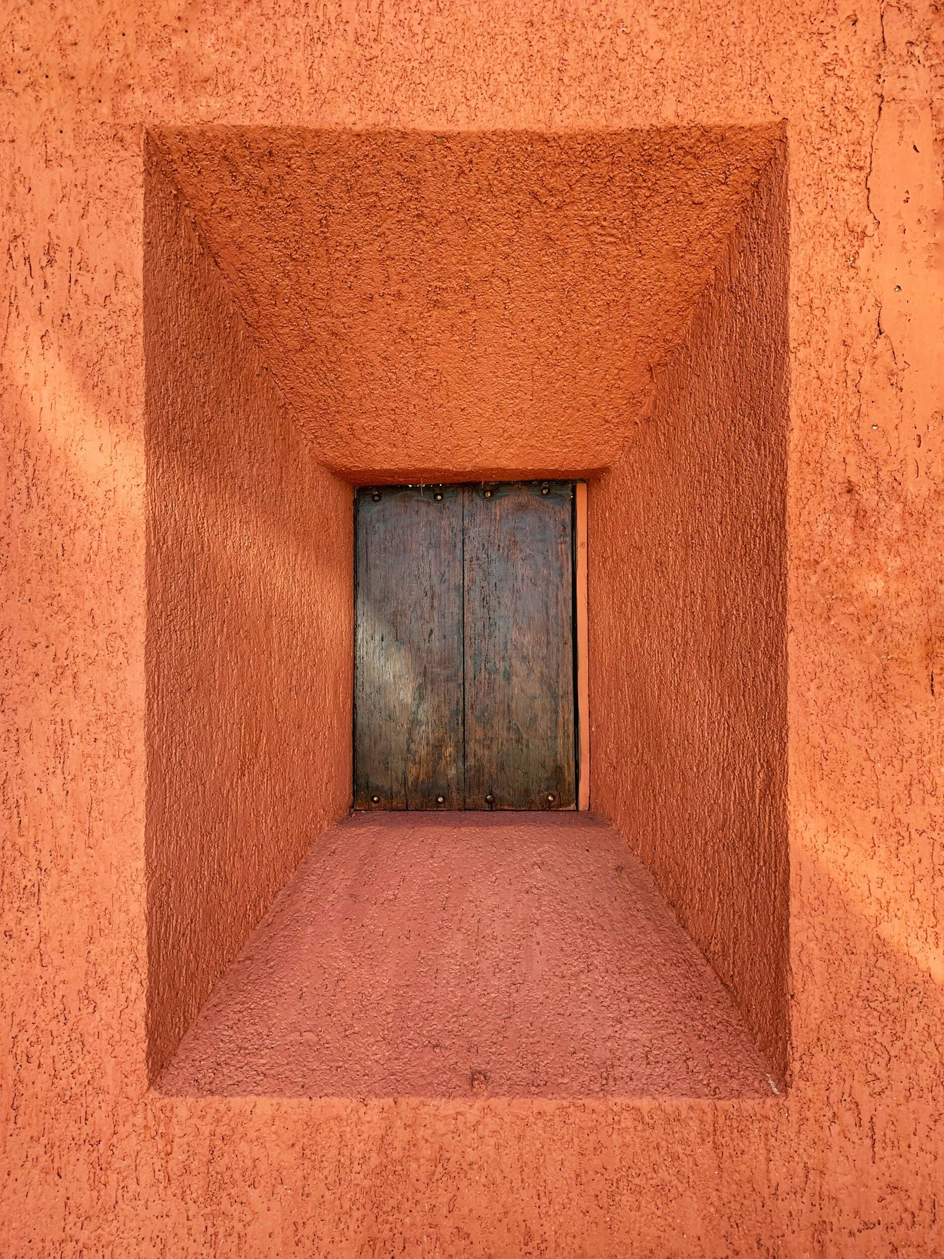 A small, rectangular window with a wooden cover set into a textured, reddish-orange wall.