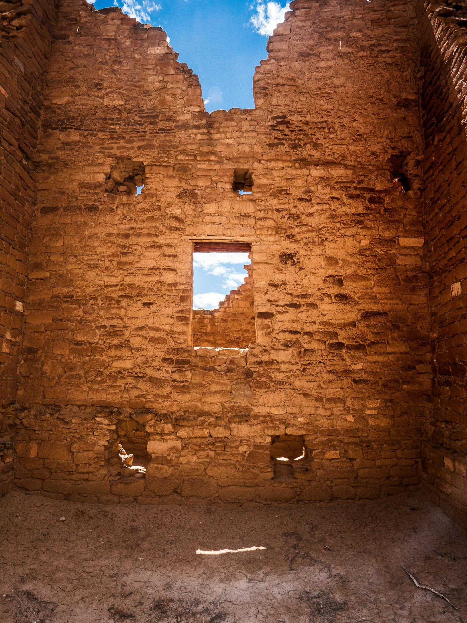 The image shows the interior of a weathered, abandoned brick building with missing portions of the wall, revealing a blue sky with scattered clouds through the openings.