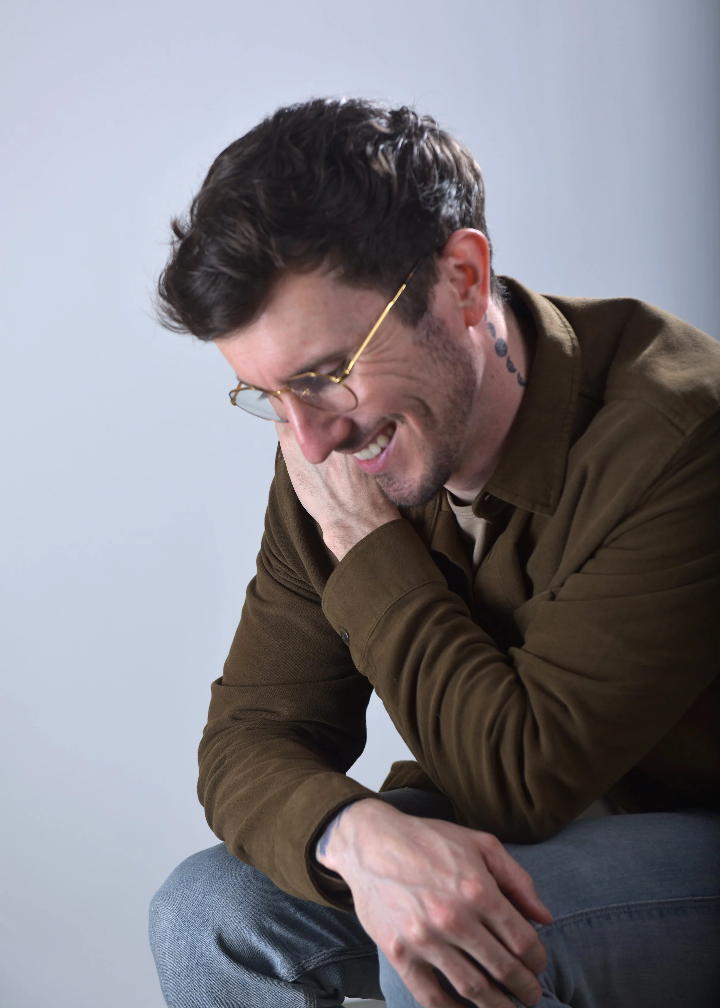 A man with dark hair, glasses, and a tattoo on his neck, smiling and looking down with his hand resting on his face, wearing a brown shirt and gray pants.