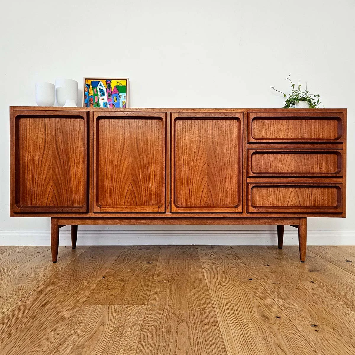 Mid-century wooden sideboard with four doors and three drawers on the right, decorated with white vases, a colorful framed artwork, and a potted plant on top, against a plain white wall.