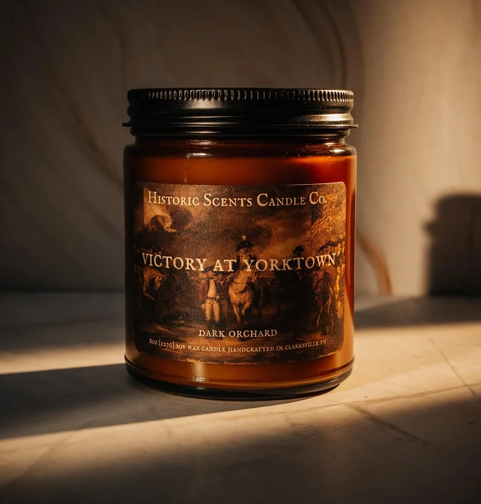 A brown jar candle labeled 'Victory at Yorktown' from Historic Scents Candle Co., depicting a historical scene, placed on a wooden surface with sunlight casting shadows.