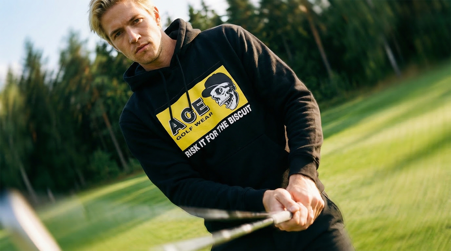 A young man with blond hair wearing a black hoodie with yellow and white graphic design, holding a golf club on a golf course.