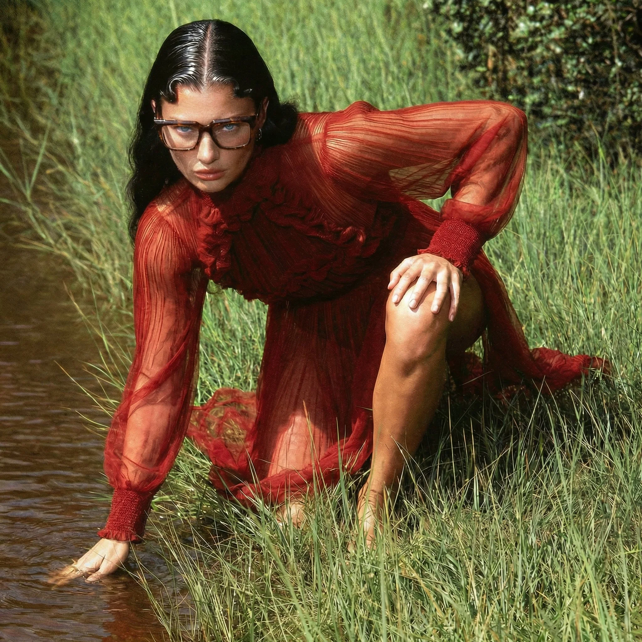 A person with black hair and glasses crouching on grass and water, wearing a red textured outfit.